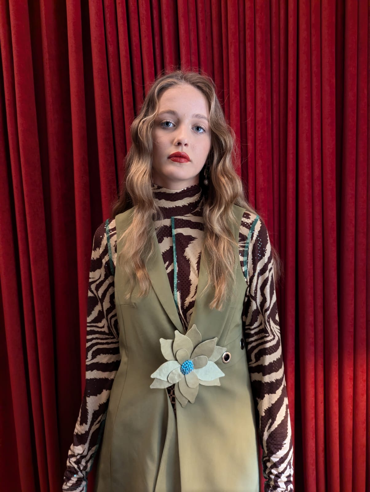 A model stands in front of a red velvet curtain wearing a unique outfit consisting of a beige vest with a large Broochella ninfea flower brooch at the waist, layered over a brown and beige zebra-print mock-neck top.