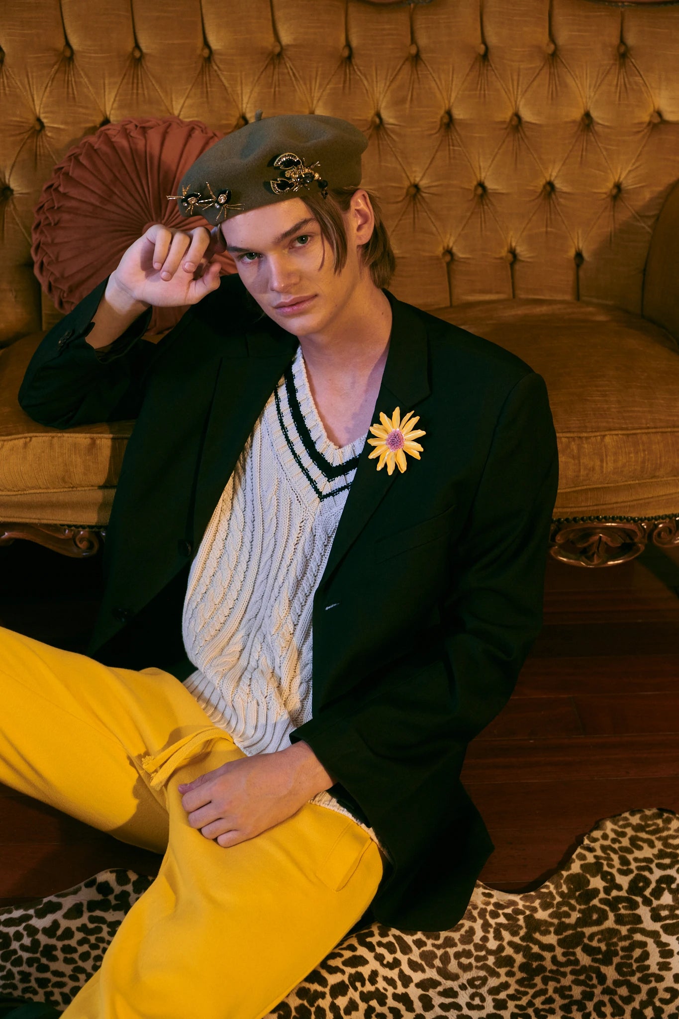 A model wearing a dark olive beret with gold star embellishments, a black blazer, and a cream knit sweater vest with a striped collar, seated on an ornate gold tufted armchair against a green wall. He is wearing the Broochella gold and black beaded spider brooch set on his beret.