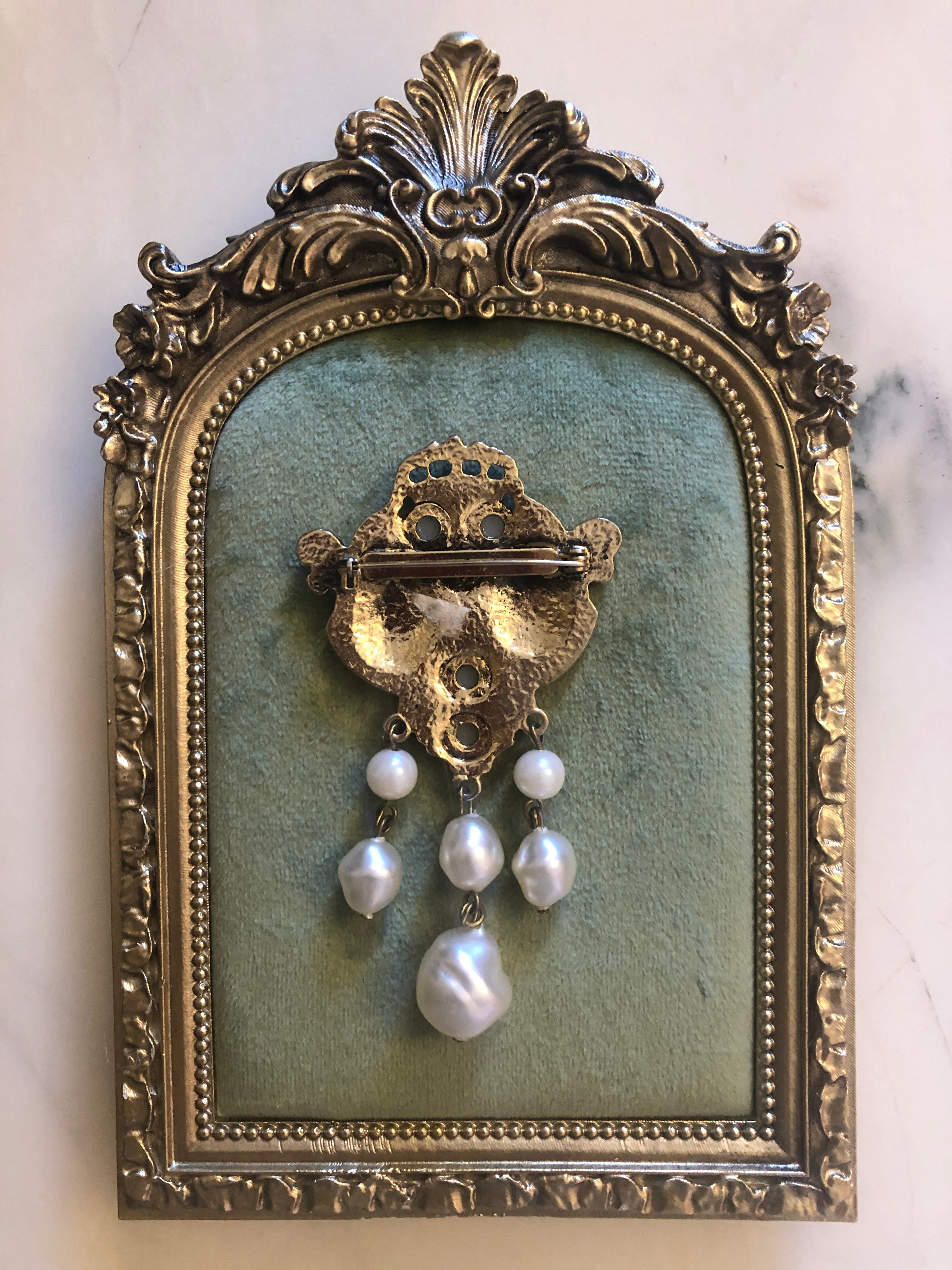 An 8cm x 5cm vintage antique gold-tone heraldic crest brooch featuring a crowned shield centrepiece with engraved coat of arms medallions, creamy seed pearl accents, ornate scrollwork cartouches, and three cascading baroque faux pearl drops, photographed on sage green velvet.
