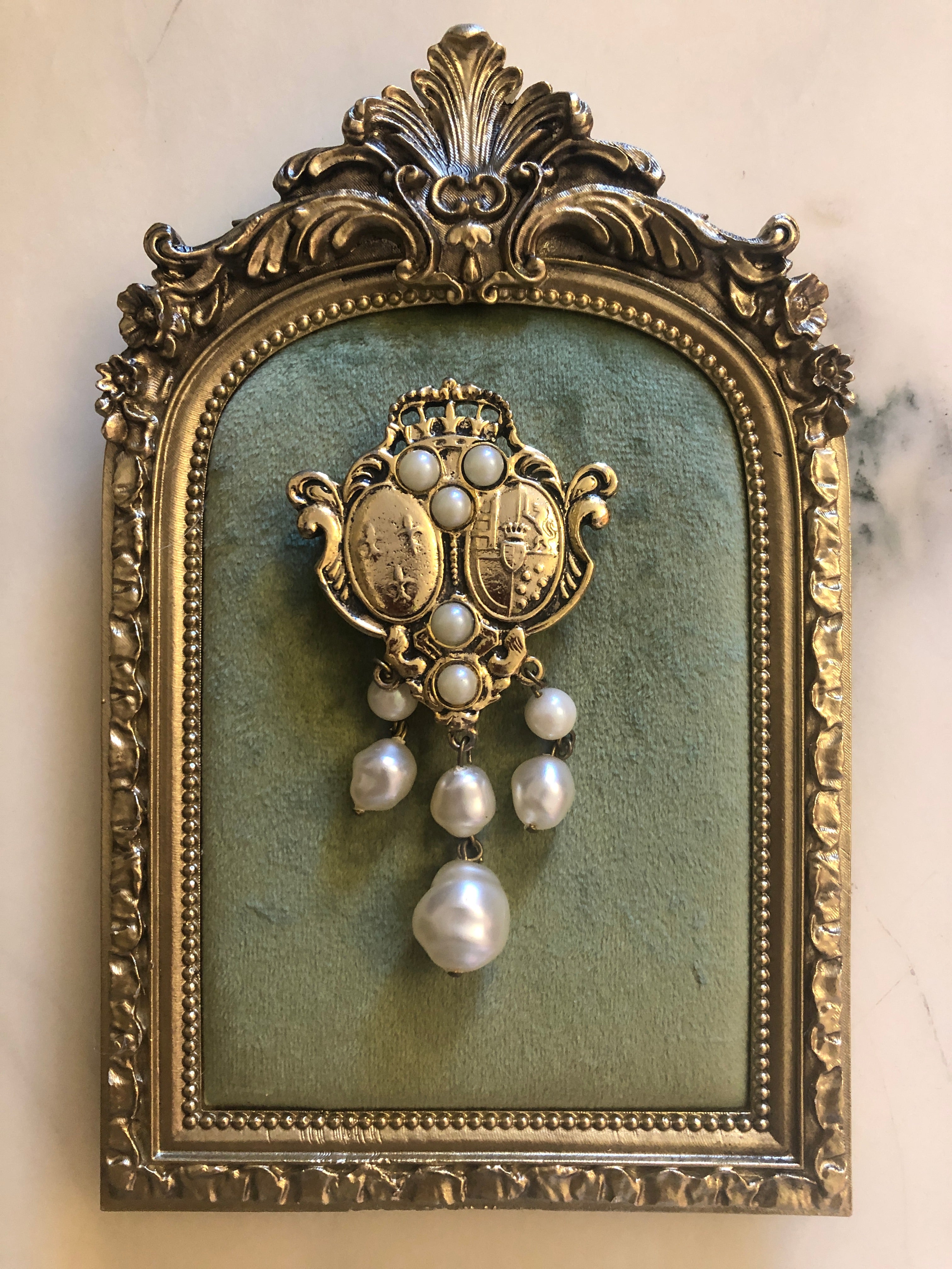 An 8cm x 5cm vintage antique gold-tone heraldic crest brooch featuring a crowned shield centrepiece with engraved coat of arms medallions, creamy seed pearl accents, ornate scrollwork cartouches, and three cascading baroque faux pearl drops, photographed on sage green velvet.