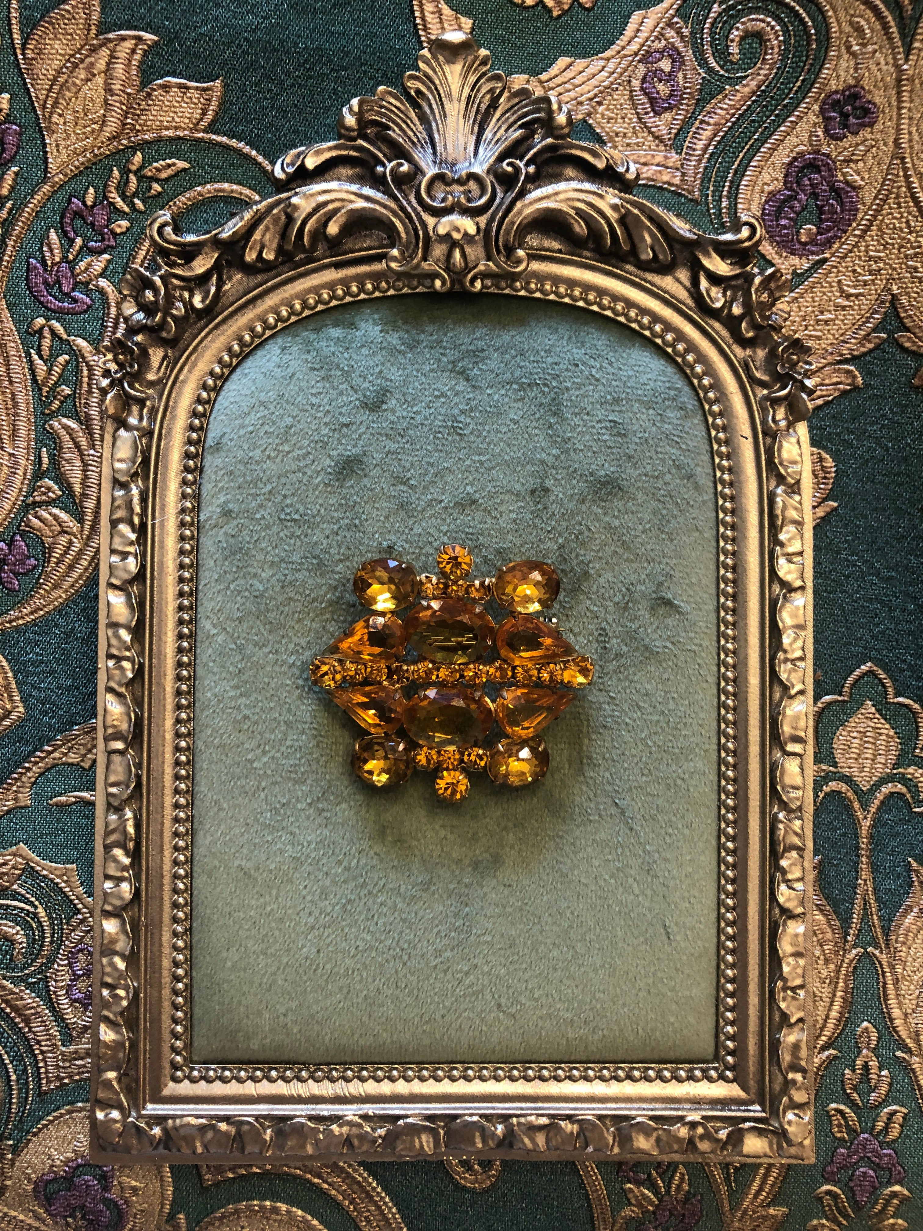 A 4.5cm x 4.5cm vintage gold-tone cluster brooch featuring large faceted oval and pear-cut amber crystal stones with graduating round amber rhinestone accents in a layered sculptural arrangement, photographed on sage green velvet.