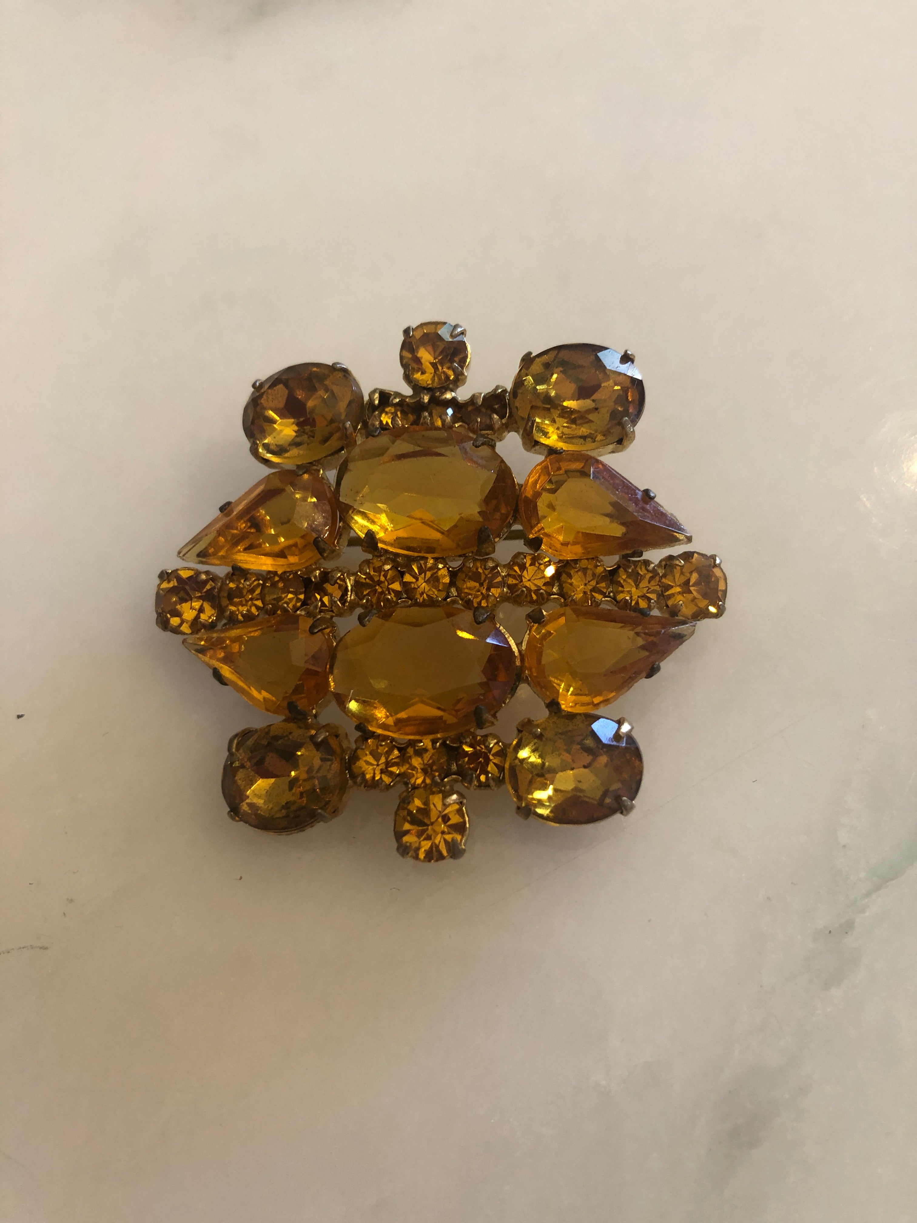 A 4.5cm x 4.5cm vintage gold-tone cluster brooch featuring large faceted oval and pear-cut amber crystal stones with graduating round amber rhinestone accents in a layered sculptural arrangement, photographed on marble table