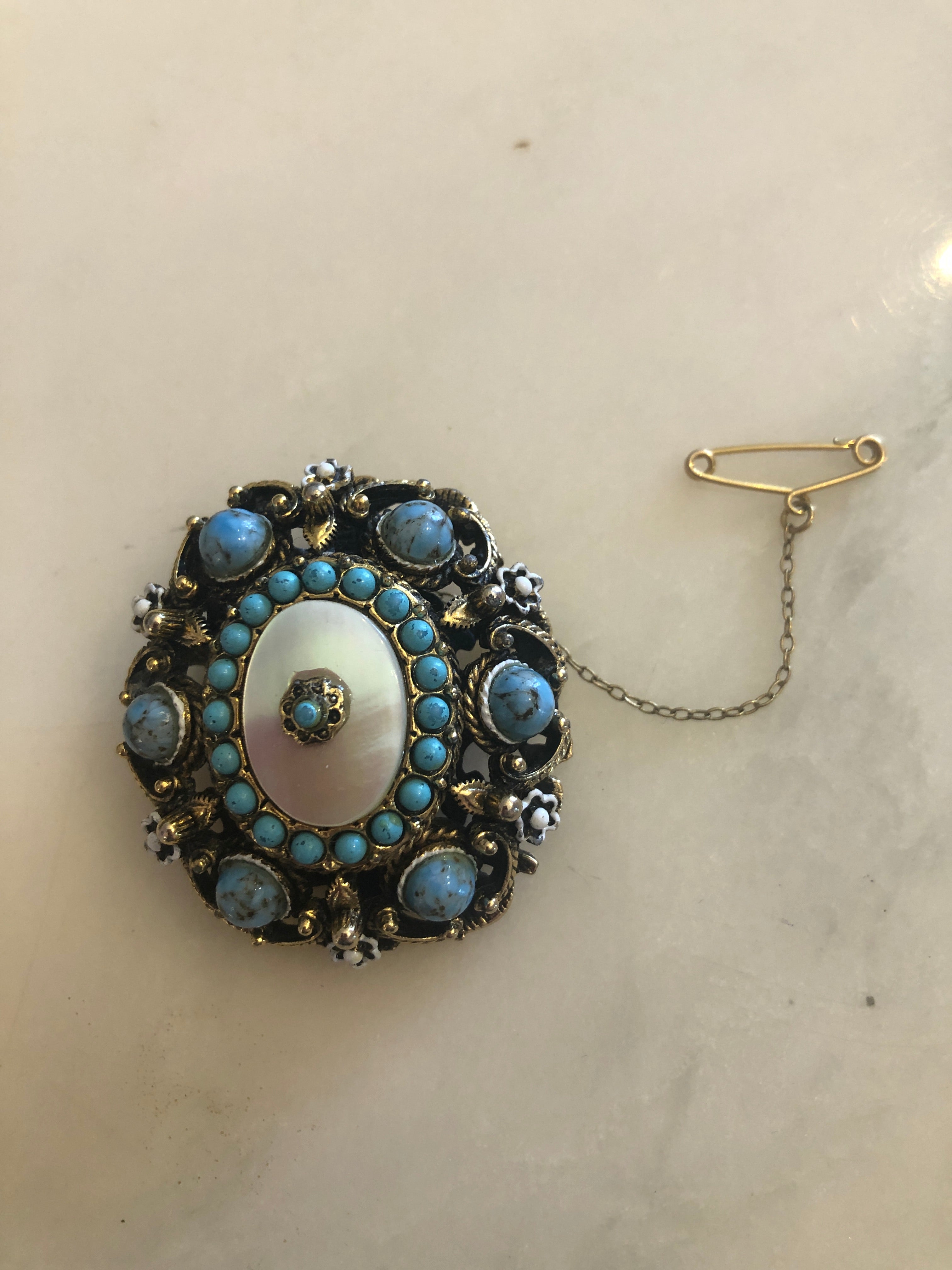 A 4cm x 4cm vintage Czech circular brooch featuring an oxidised and gold-tone openwork frame, a central mother of pearl oval cabochon surrounded by turquoise seed beads, eight blue marbled outer cabochons, white enamel flower accents, and an original gold-tone safety chain.