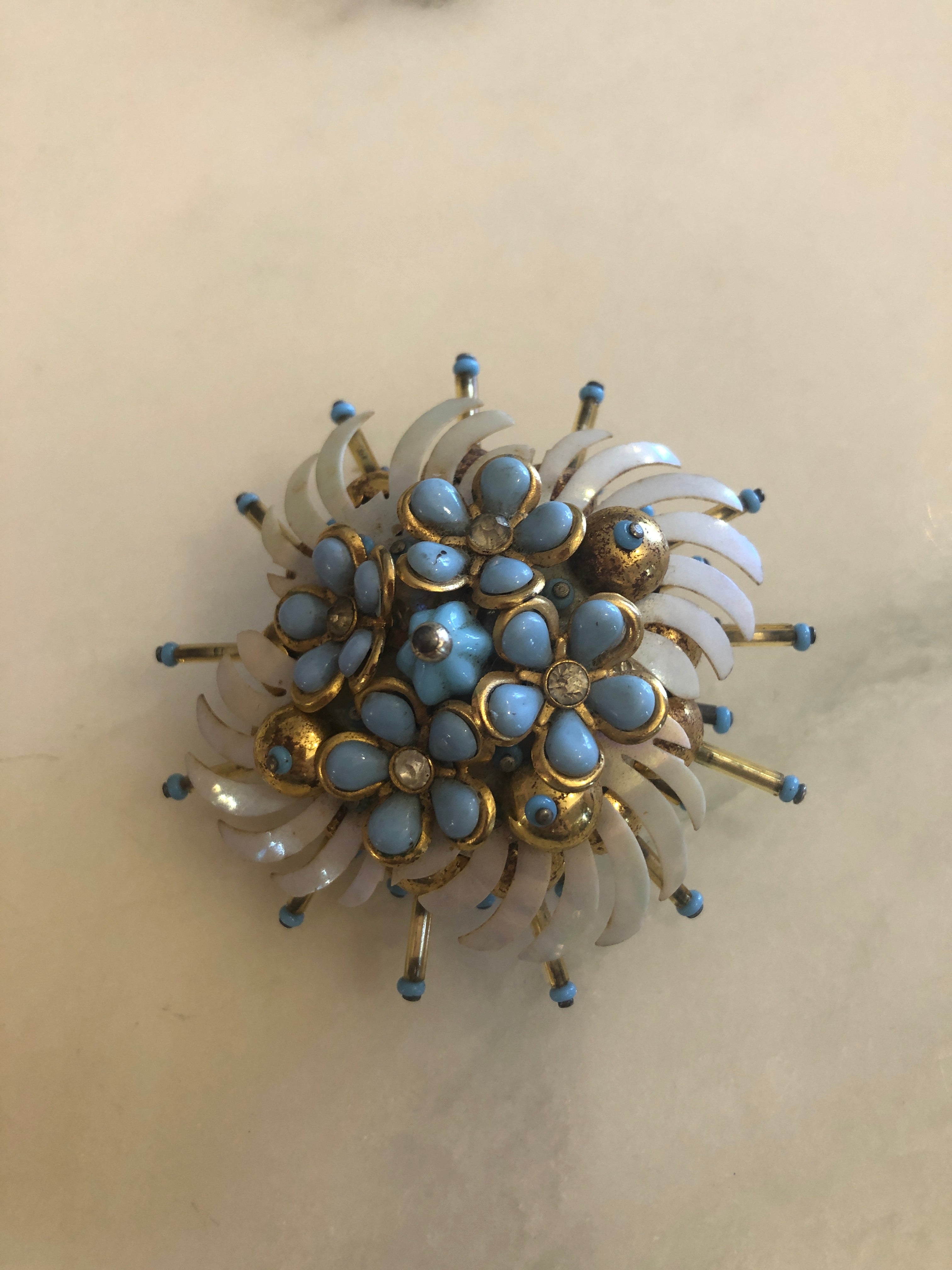 A 6cm x 6cm vintage gold-tone floral burst brooch featuring layered white enamel swept petals, a dense centre cluster of powder blue cabochon flowers set in gold-tone bezels with crystal rhinestone accents, and radiating spike pins tipped in powder blue.
