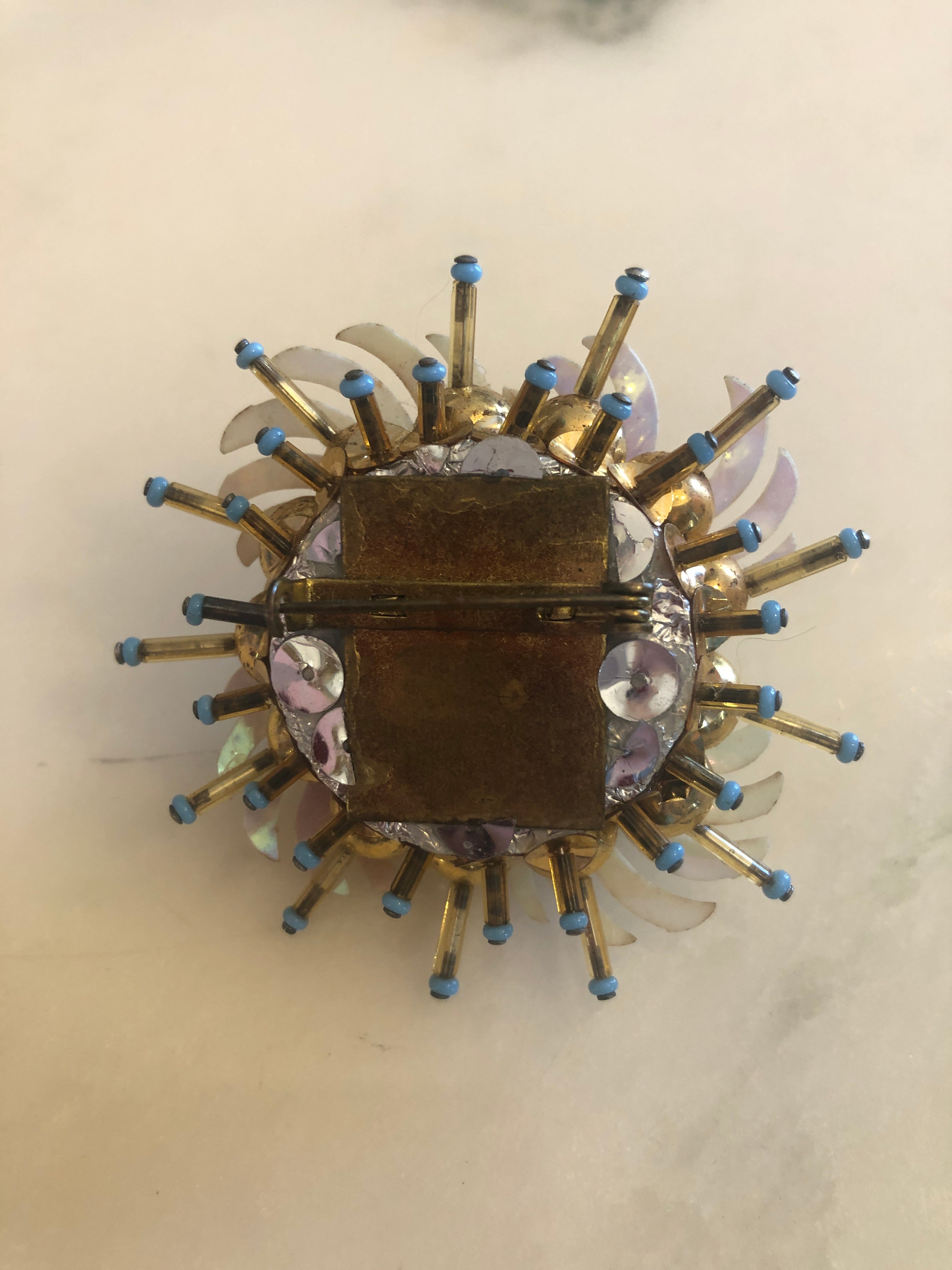 A 6cm x 6cm vintage gold-tone floral burst brooch featuring layered white enamel swept petals, a dense centre cluster of powder blue cabochon flowers set in gold-tone bezels with crystal rhinestone accents, and radiating spike pins tipped in powder blue.