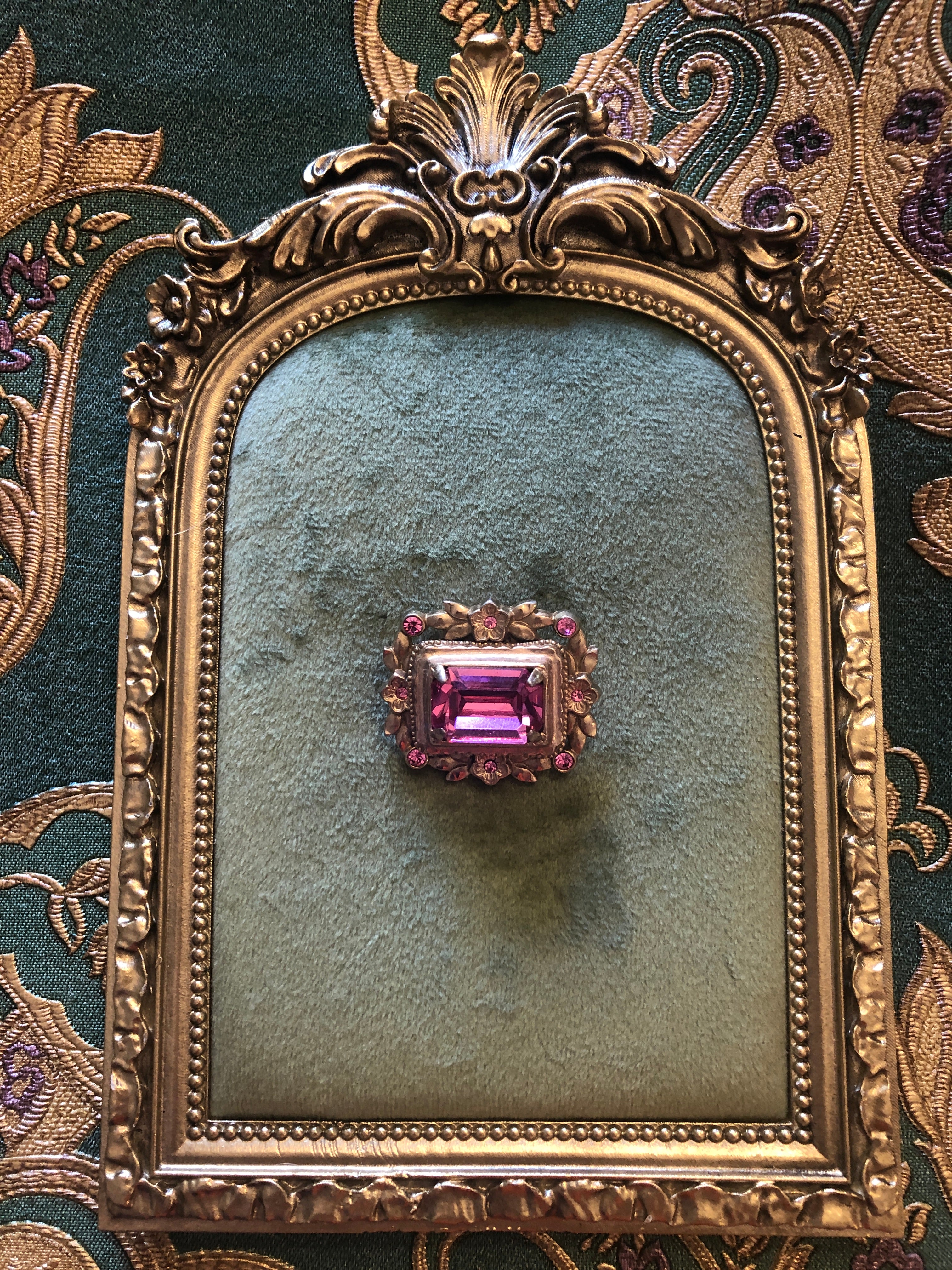 A small vintage gold-tone rectangular brooch featuring a large faceted emerald-cut pink crystal centre stone set within a beaded frame, surrounded by a floral wreath of matte gold-tone flower and leaf motifs with six pink rhinestone accents. Photographed on gold frame, with a green velvet centre. The background is green and gold damask wallpaper