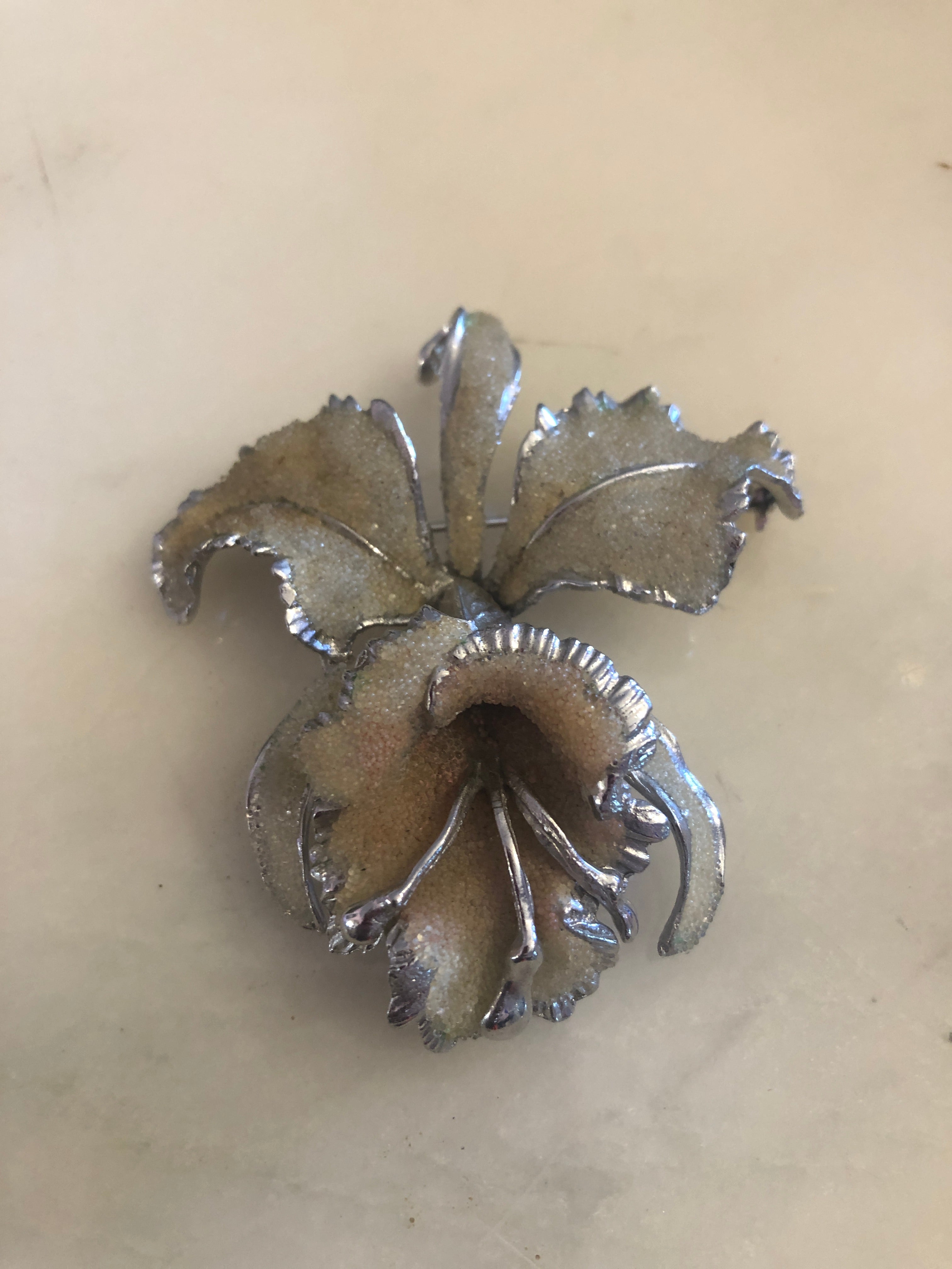 A vintage signed Elizabeth Reimer Arcansas Coralene silver-tone orchid brooch featuring five sculptural petals with a fine frosted sand-texture coralene surface, polished serrated silver-tone metal petal edges and a three-dimensional trumpet centre, photographed on a white marble surface.