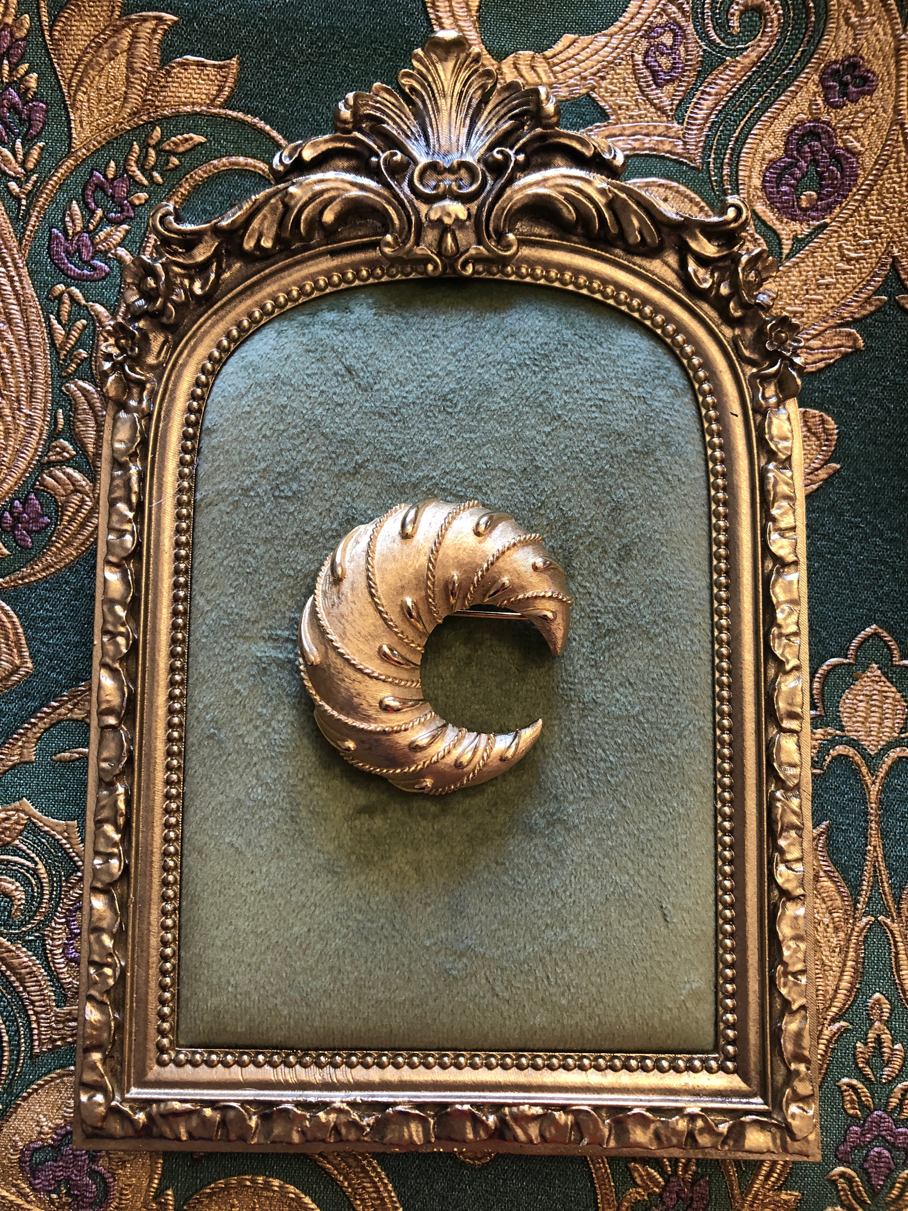A vintage Trifari gold-tone Cornetto brooch featuring a sculptural open swirl form with finely brushed crosshatch-textured surface, polished rope twist edging, raised teardrop dot accents and layered ribbed detailing throughout the curved crescent body, photographed on a green velvet and gold frame. The background is green and gold damask wallpaper