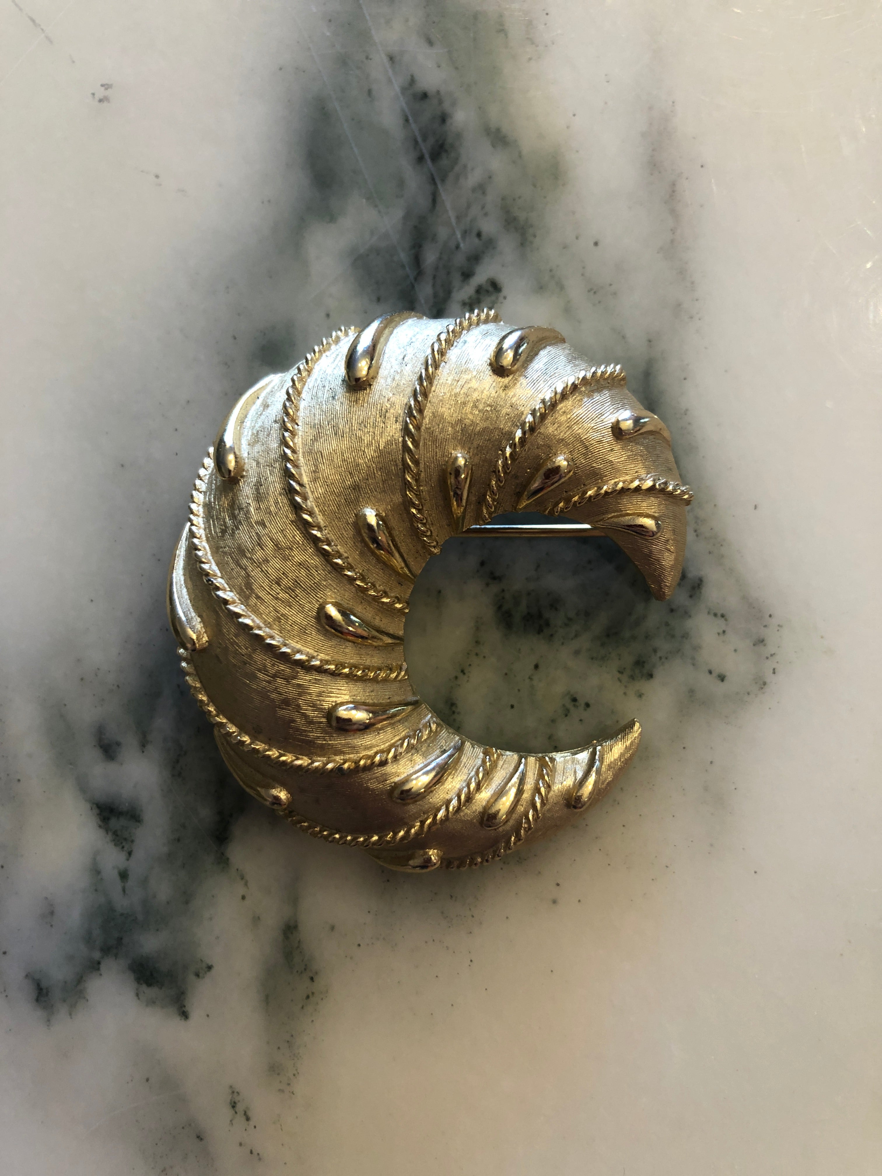 A vintage Trifari gold-tone Cornetto brooch featuring a sculptural open swirl form with finely brushed crosshatch-textured surface, polished rope twist edging, raised teardrop dot accents and layered ribbed detailing throughout the curved crescent body, photographed on a white marble surface.
