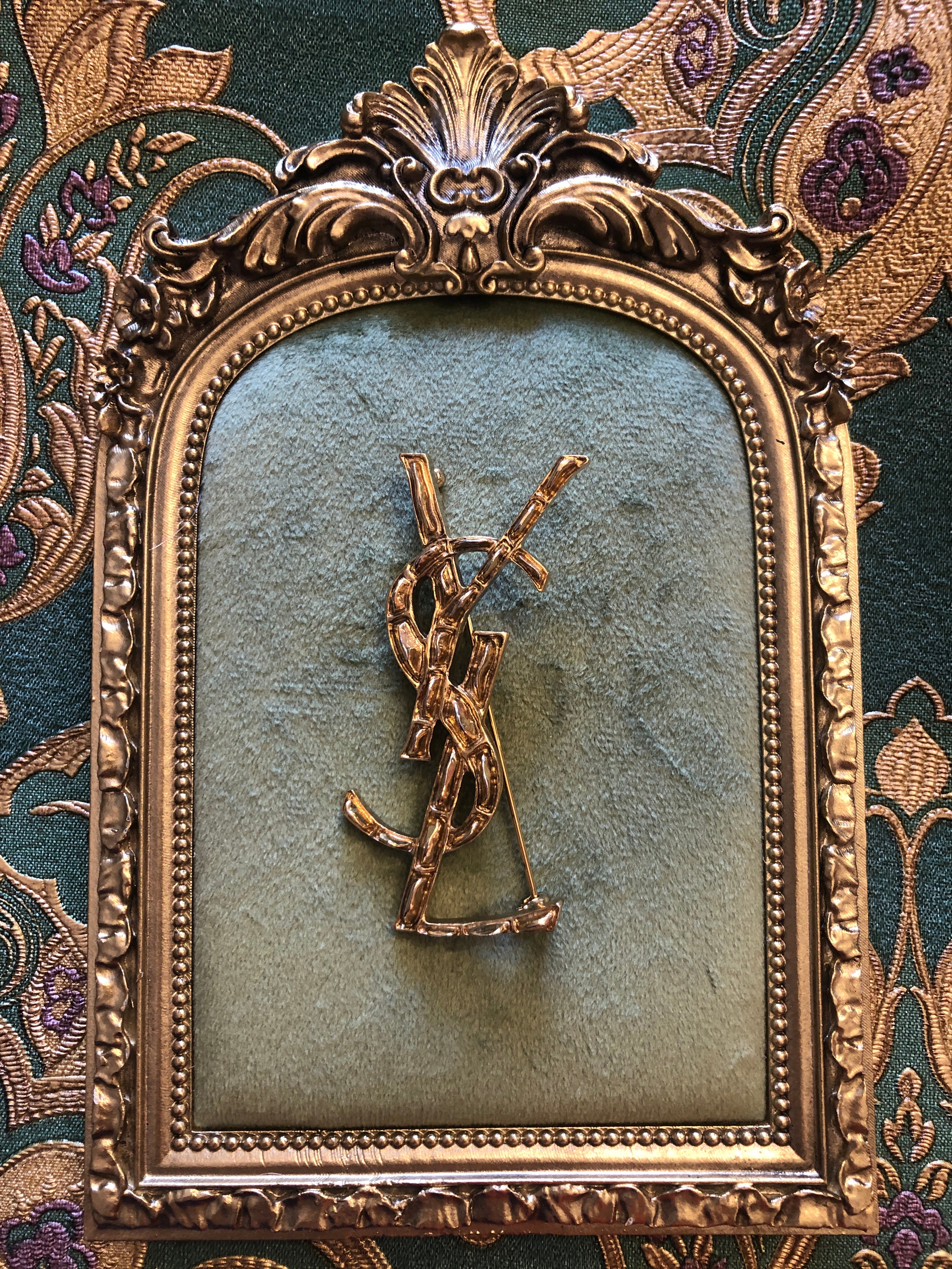 A vintage Yves Saint Laurent gold-tone logo brooch featuring the iconic interlocking YSL monogram in a large sculptural bamboo-effect textured format with polished raised edges, photographed on a green velvet and gold framed tray. The background is gerrn and gold damask wallpaper