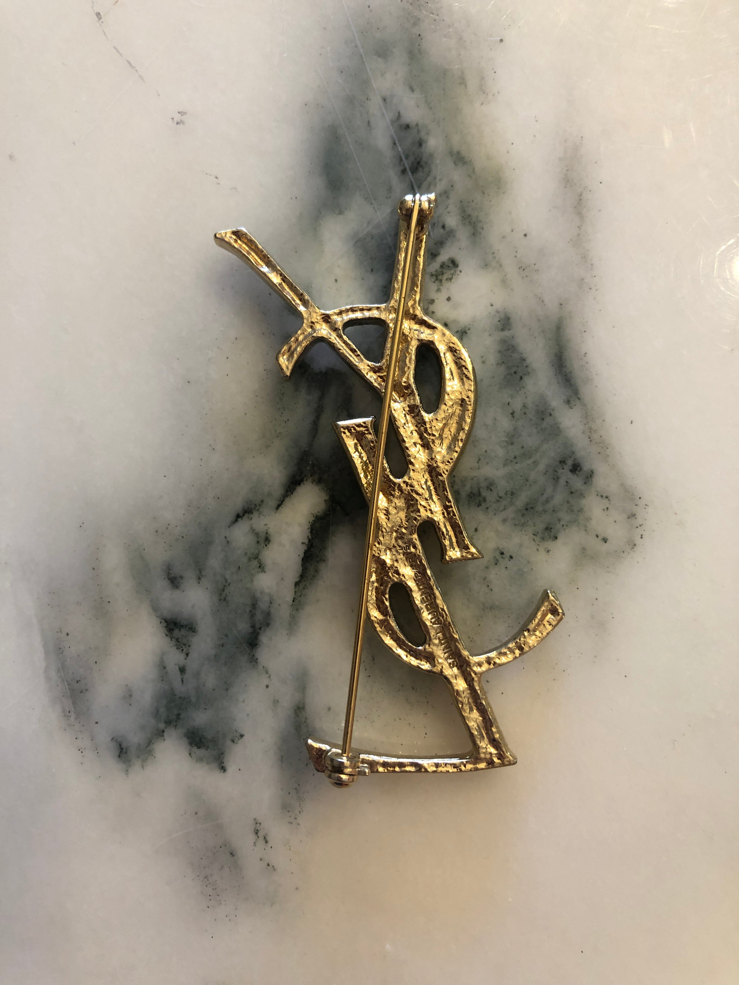 A vintage Yves Saint Laurent gold-tone logo brooch featuring the iconic interlocking YSL monogram in a large sculptural bamboo-effect textured format with polished raised edges, photographed on a white marble surface.