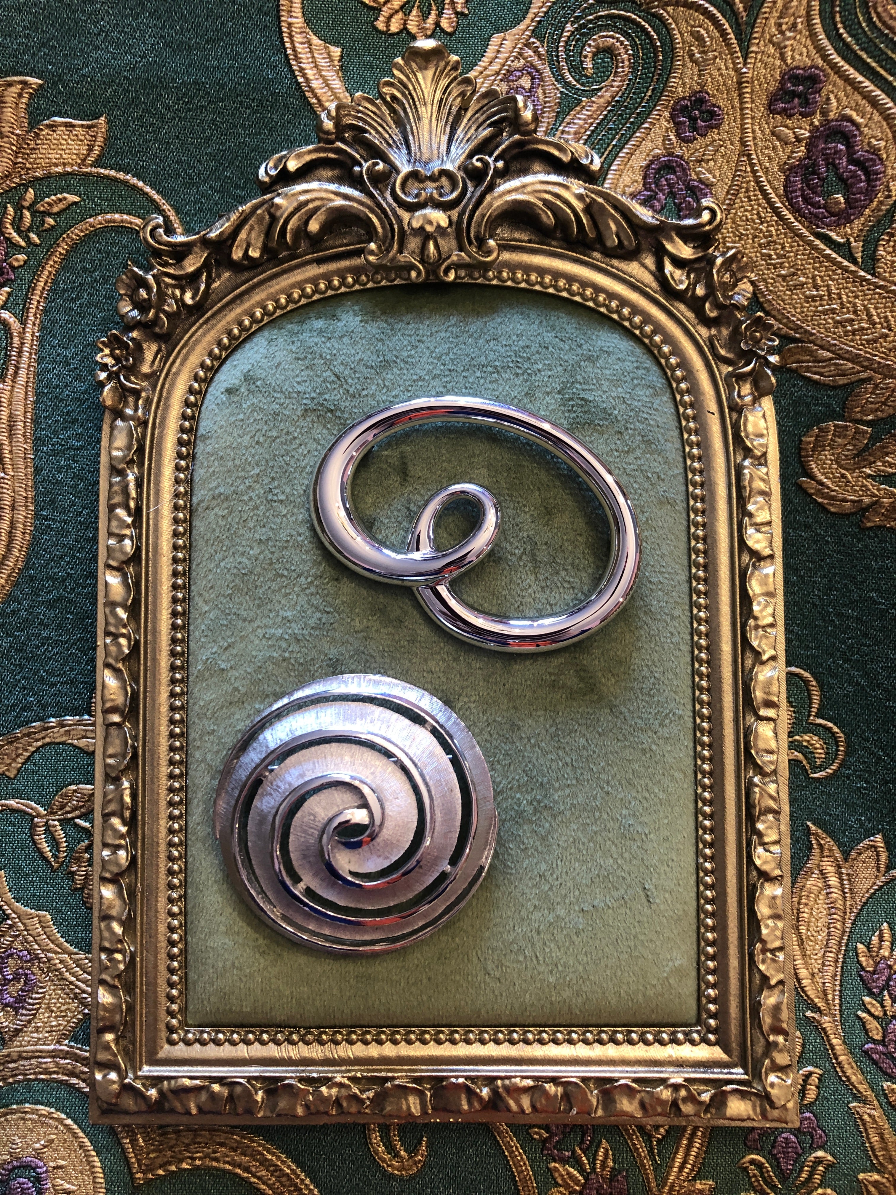 Two vintage silver-tone brooches photographed together on a green velvet tray. The background us green and gold damake wallpaper Top: a Trifari polished silver-tone open loop brooch featuring a fluid cursive swirl form with a small inner loop detail. Bottom: a Monet circular silver-tone brooch with a brushed crosshatch-textured surface and a polished inset spiral motif with a raised outer rim