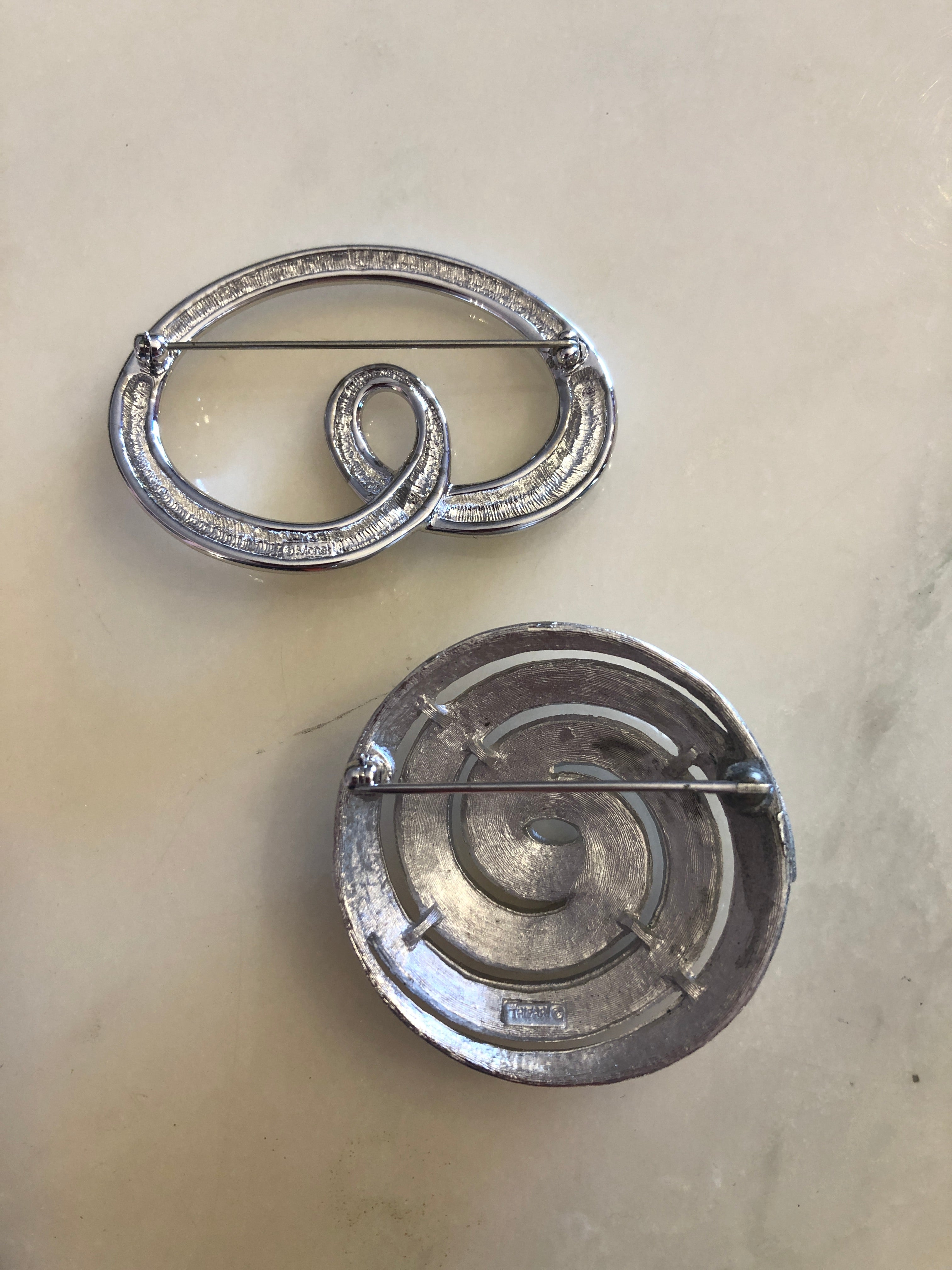 Two vintage silver-tone brooches photographed together on a white marble surface. Top: a Trifari polished silver-tone open loop brooch featuring a fluid cursive swirl form with a small inner loop detail. Bottom: a Monet circular silver-tone brooch with a brushed crosshatch-textured surface and a polished inset spiral motif with a raised outer rim.