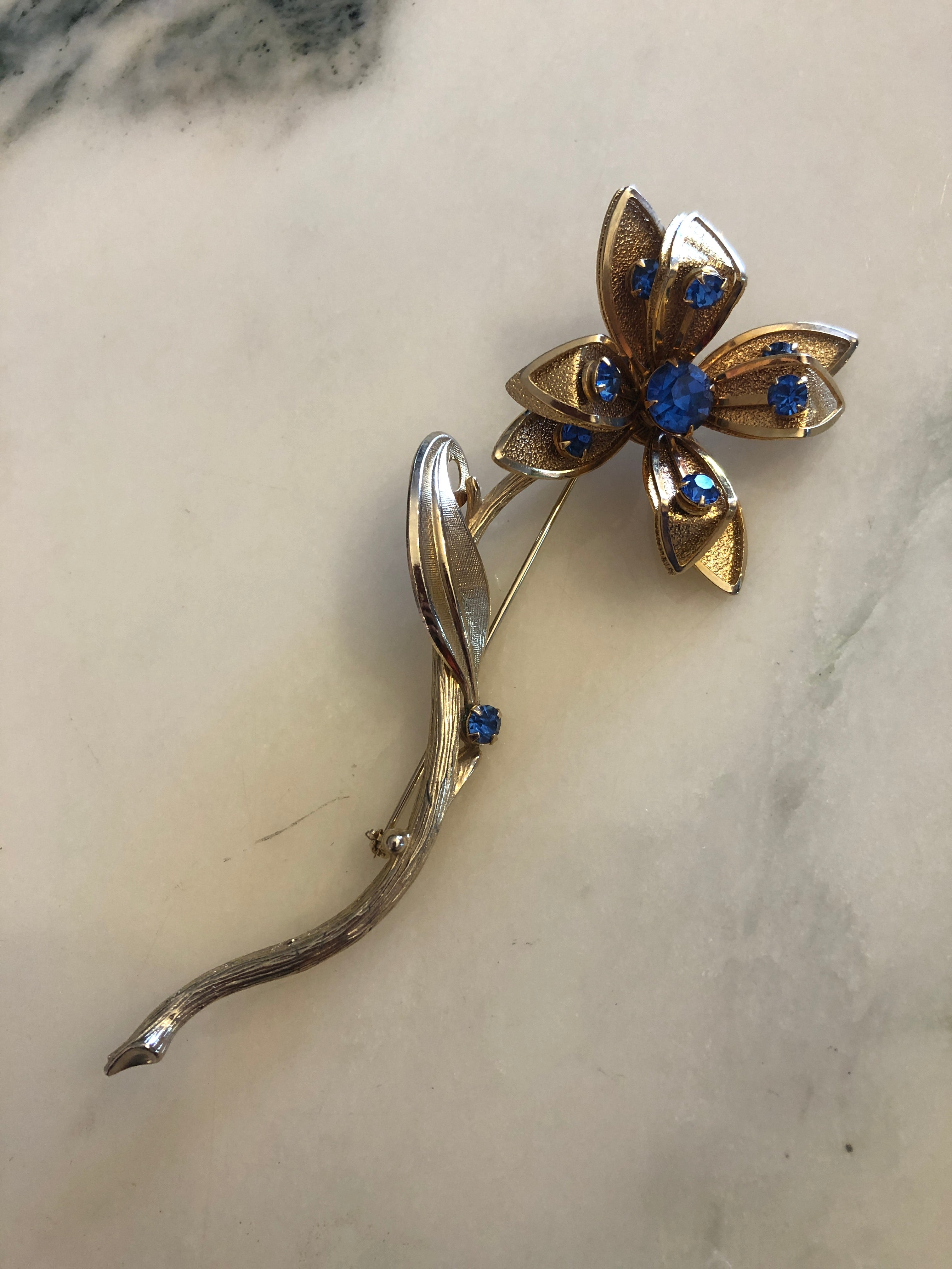 A large vintage gold and silver-tone Fiore Azzurro flower stem brooch featuring five textured gold-tone petals with polished silver-tone tips set with sapphire blue round crystal rhinestones, a central sapphire blue rhinestone cluster, a single silver-tone leaf accent and an additional sapphire blue stone on the elongated curved textured stem, photographed on a white marble surface.