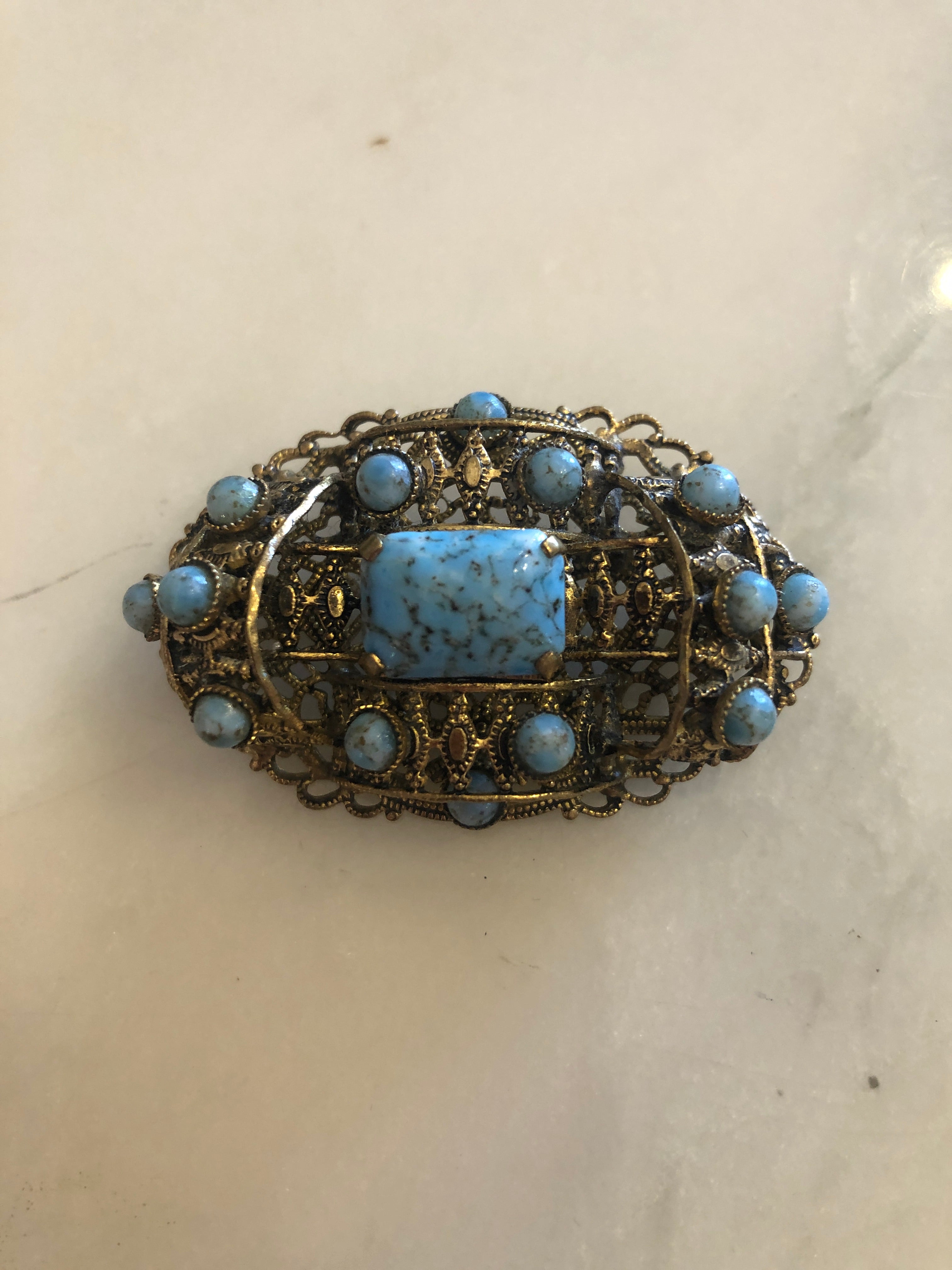 A vintage Czech Tesoro Azzurro gold-tone oval filigree brooch featuring a square turquoise marbled cabochon centre stone set within an intricate openwork granulated scrollwork frame, surrounded by fourteen round turquoise marbled cabochon stones set along the outer border, photographed on a white marble surface.