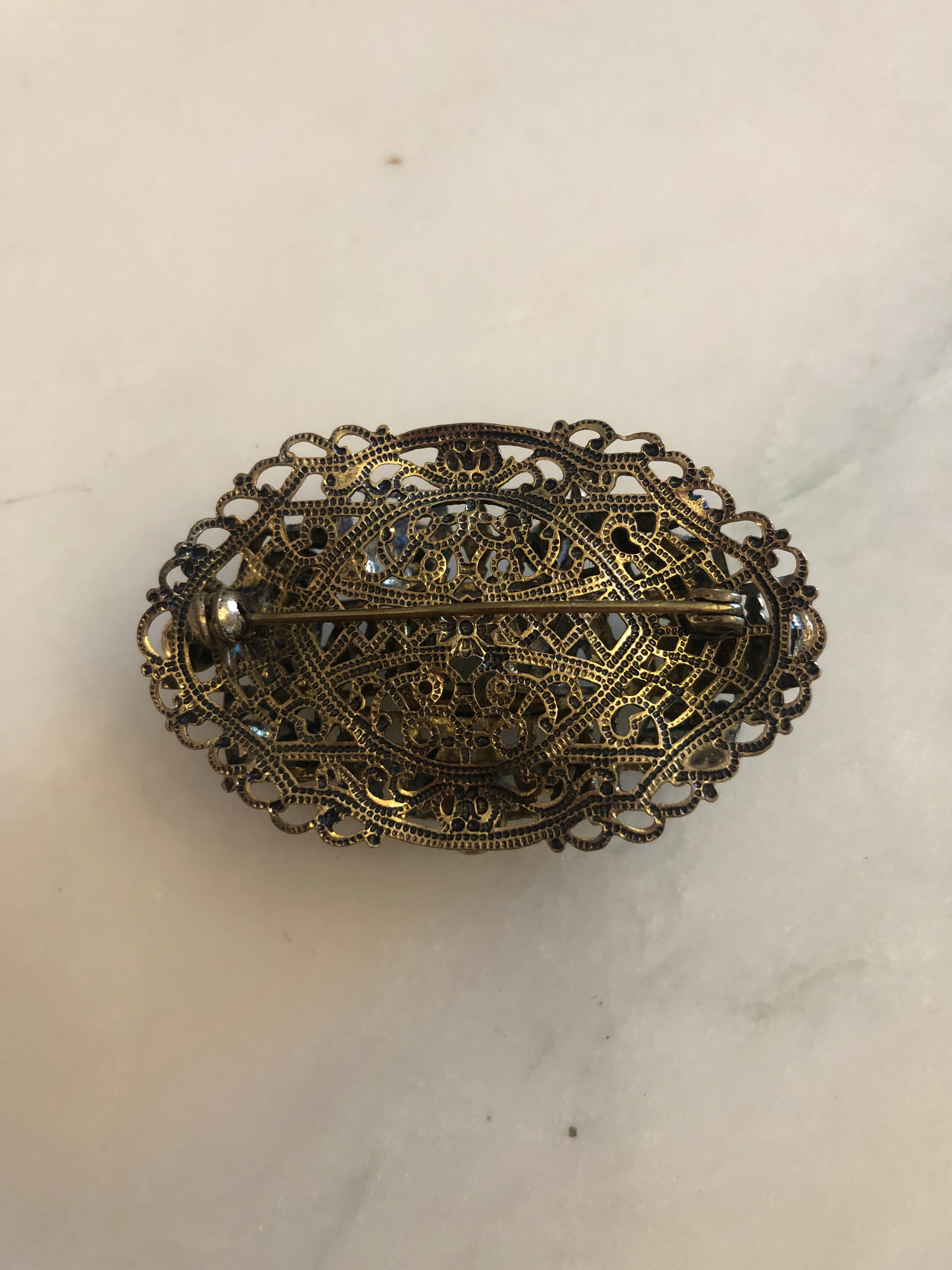 A vintage Czech Tesoro Azzurro gold-tone oval filigree brooch featuring a square turquoise marbled cabochon centre stone set within an intricate openwork granulated scrollwork frame, surrounded by fourteen round turquoise marbled cabochon stones set along the outer border, photographed on a white marble surface.