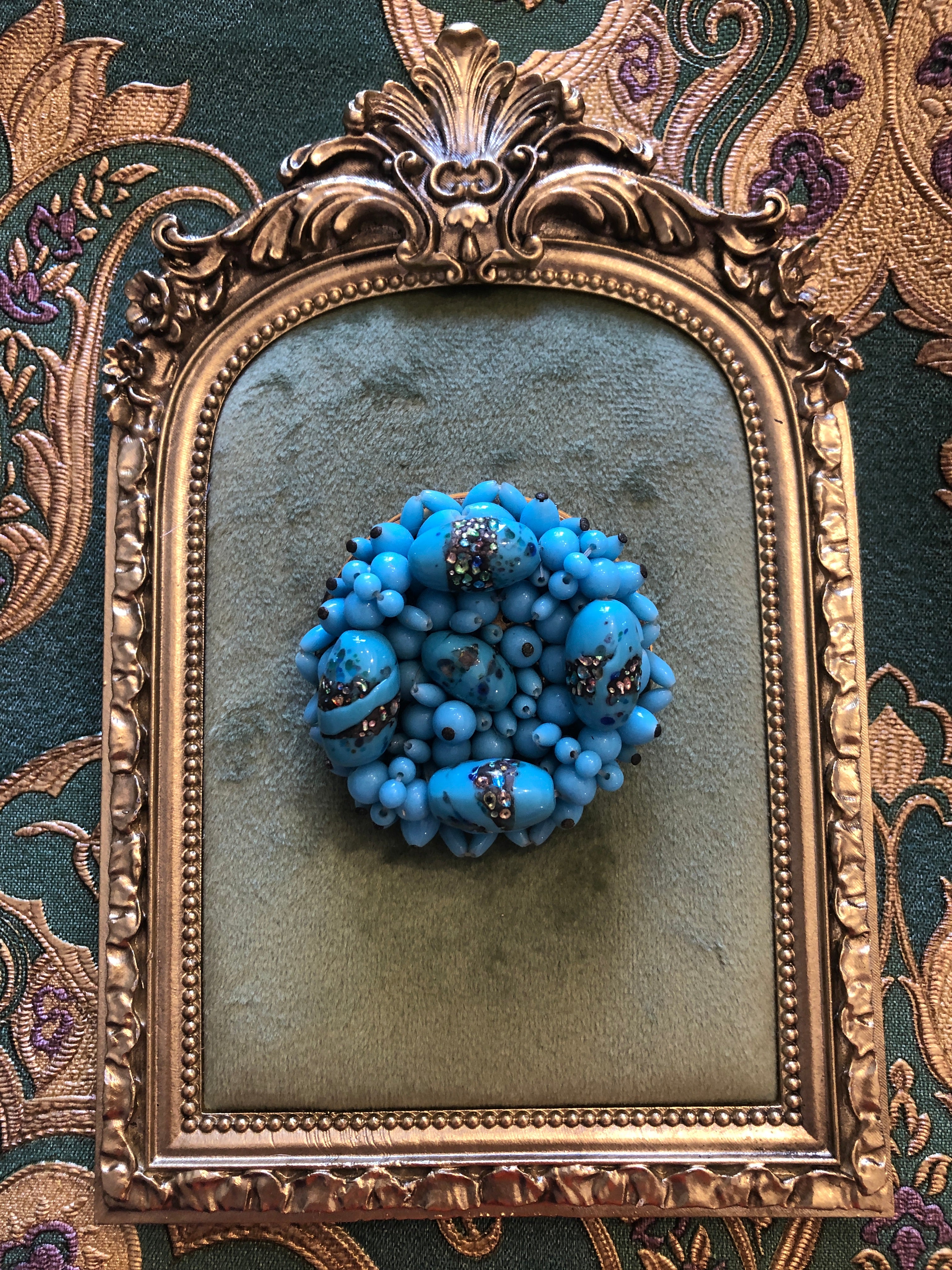 A vintage Laguna Turchese circular cluster brooch featuring a dense domed arrangement of graduating powder blue glass beads in various sizes, interspersed with four larger oval focal beads decorated with aurora borealis crystal rhinestone and metallic foil encrusted band details, photographed on a green velvet tray with a gold frame. The background is gold and green damask wallpaper