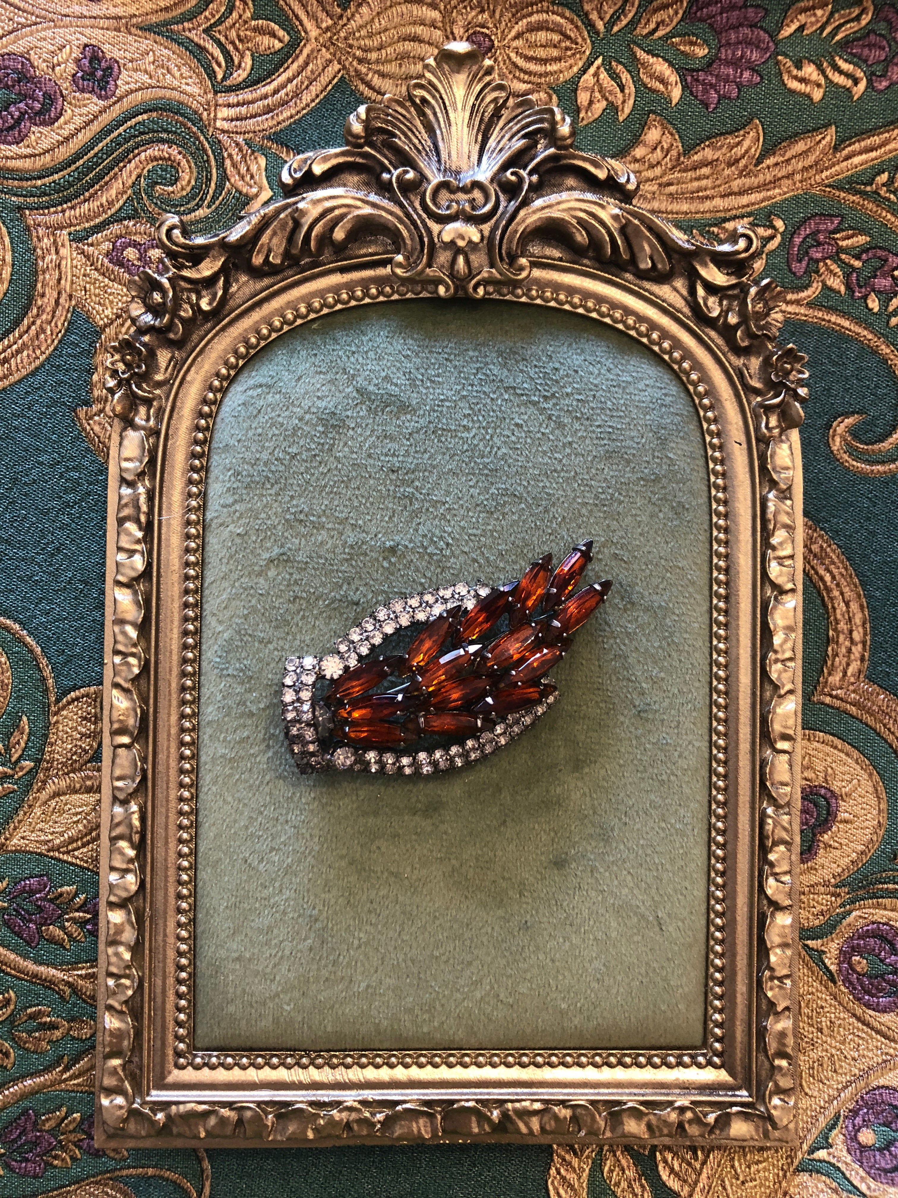 A 3cm x 6cm vintage dark silver-tone flame-shaped brooch with layered deep amber navette-cut crystals bordered by a curved double row of clear round crystal rhinestones. Photographed on a green velvet tray, with an ornate, gold frame.  The background is green and gold damask wallpaper