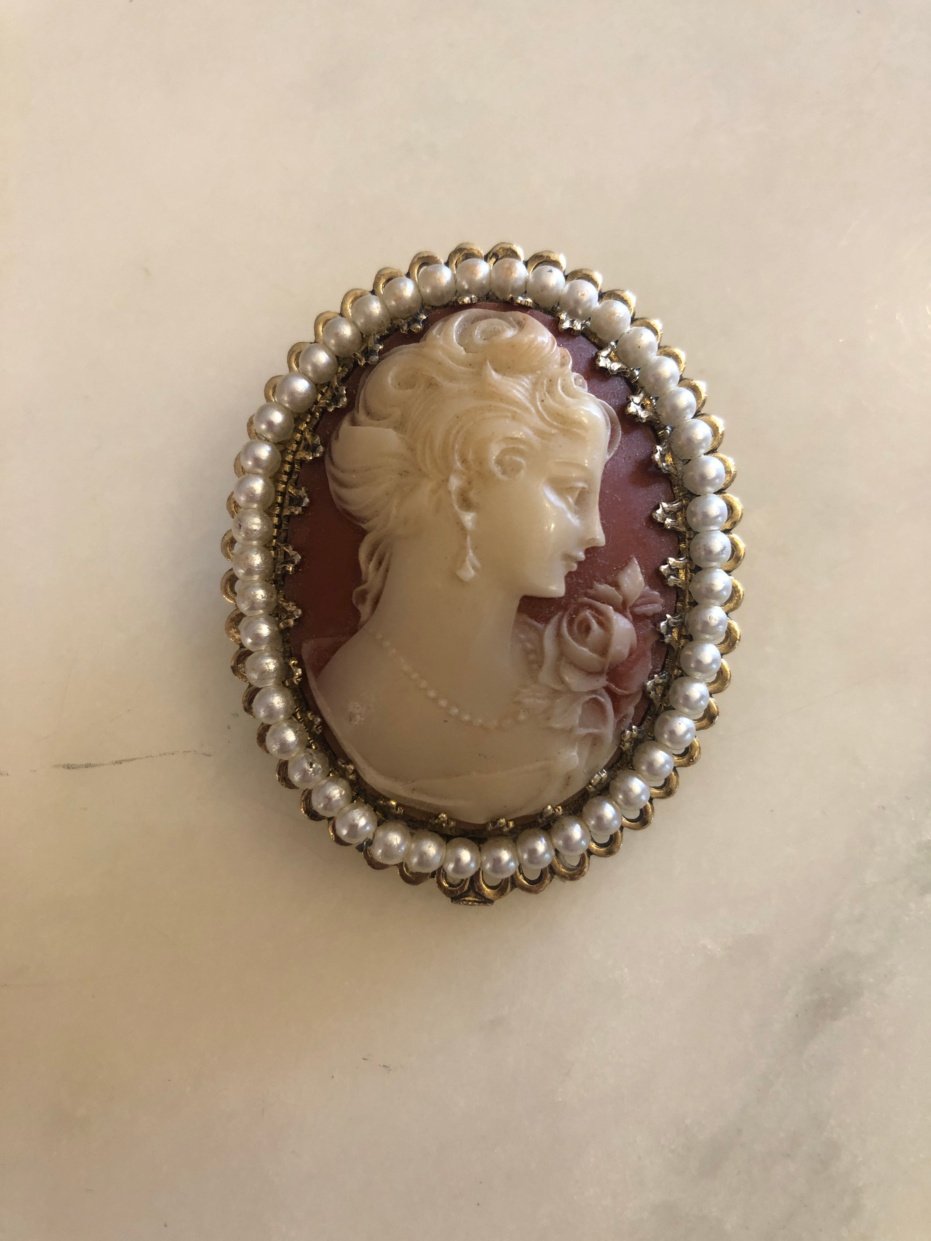 A 5.5cm x 4cm vintage shell cameo brooch with a carved cream portrait of a lady with upswept curls, drop earring and rose detail on a blush pink ground, set in a gold-tone filigree frame with a full seed pearl border, photographed on a white marble surface.