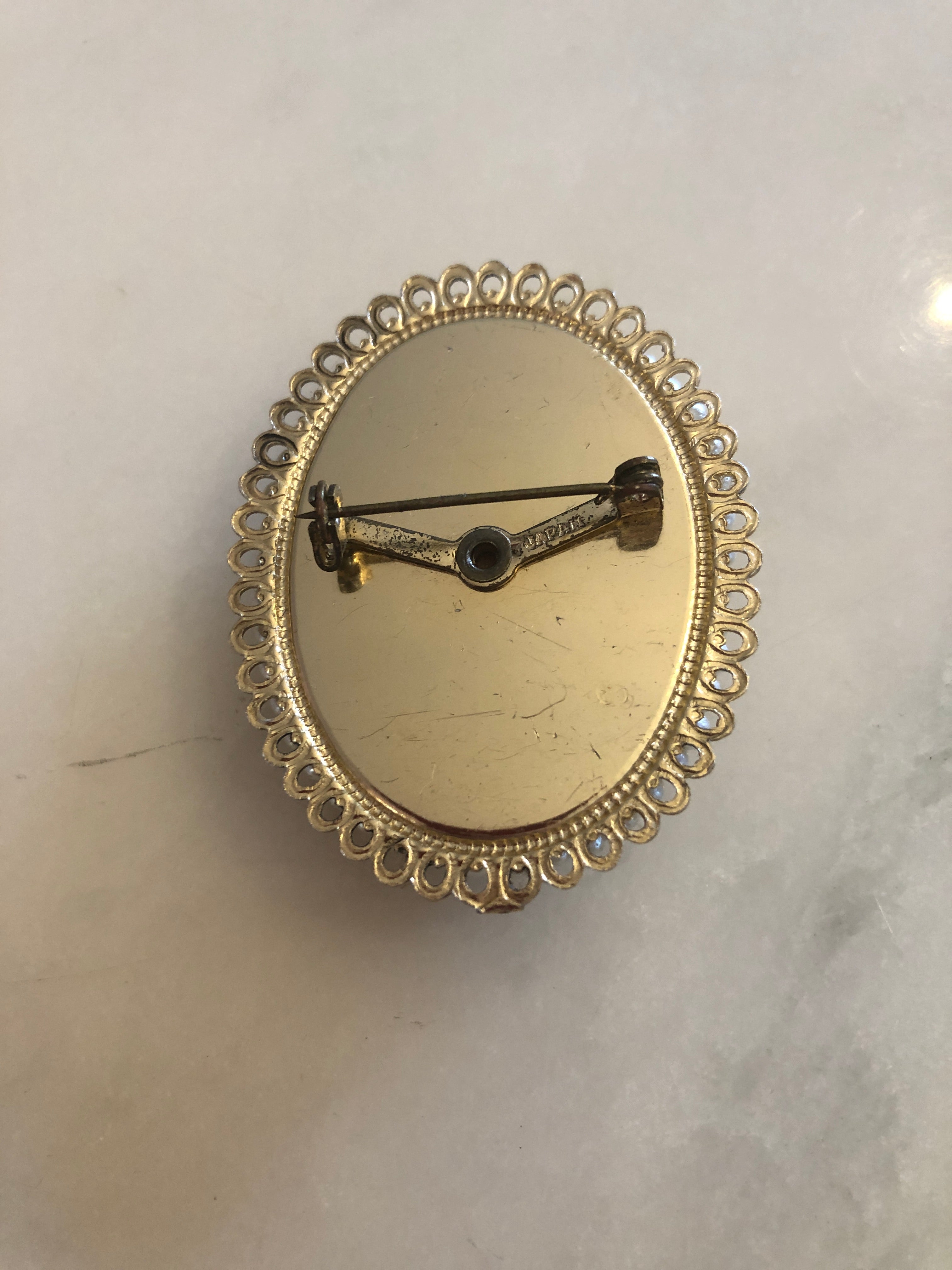 A 5.5cm x 4cm vintage shell cameo brooch with a carved cream portrait of a lady with upswept curls, drop earring and rose detail on a blush pink ground, set in a gold-tone filigree frame with a full seed pearl border, photographed on a white marble surface.