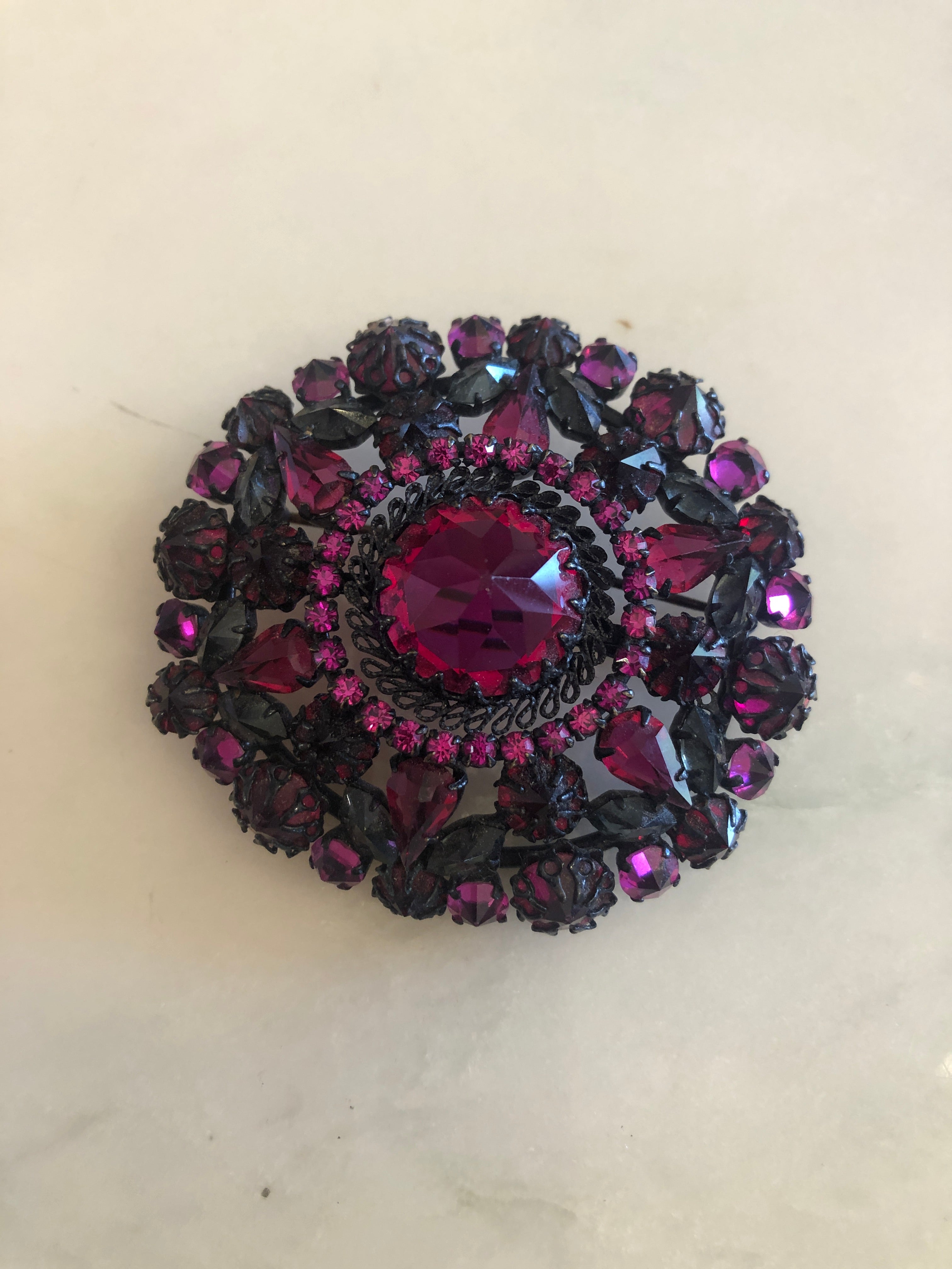 A 6cm x 6cm vintage oxidised black-tone circular brooch with a large faceted ruby red oval centre stone surrounded by layered rings of hot magenta and purple pear-cut and round crystal rhinestones, photographed on a white marble surface.