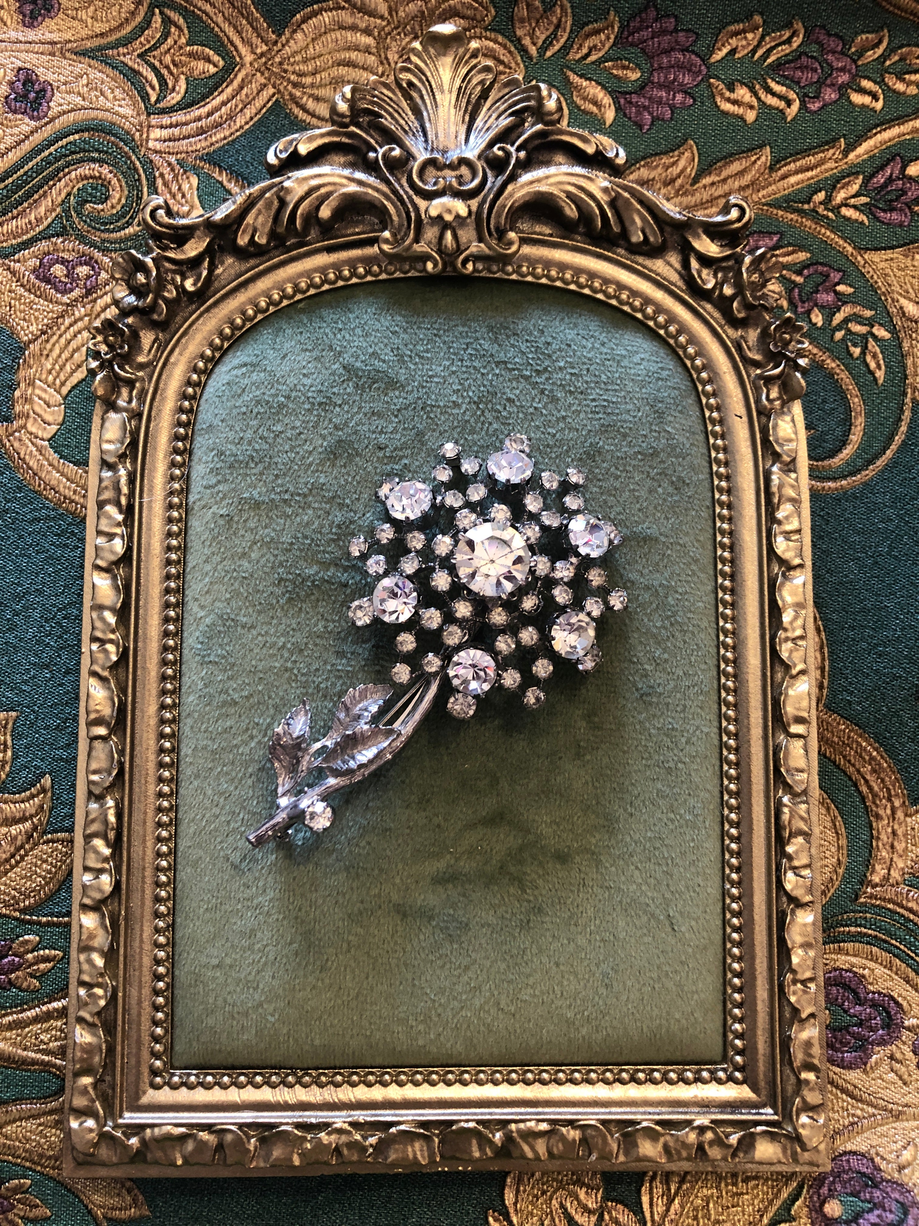 A 7.5cm x 4.5cm vintage silver-tone crystal floral spray brooch with a graduating clear round crystal rhinestone flower head on fine wire stems and a textured silver-tone leaf and stem base. Photographed on a green velvet tray, with an ornate gold frame. The background is green and gold damask wallpaper.