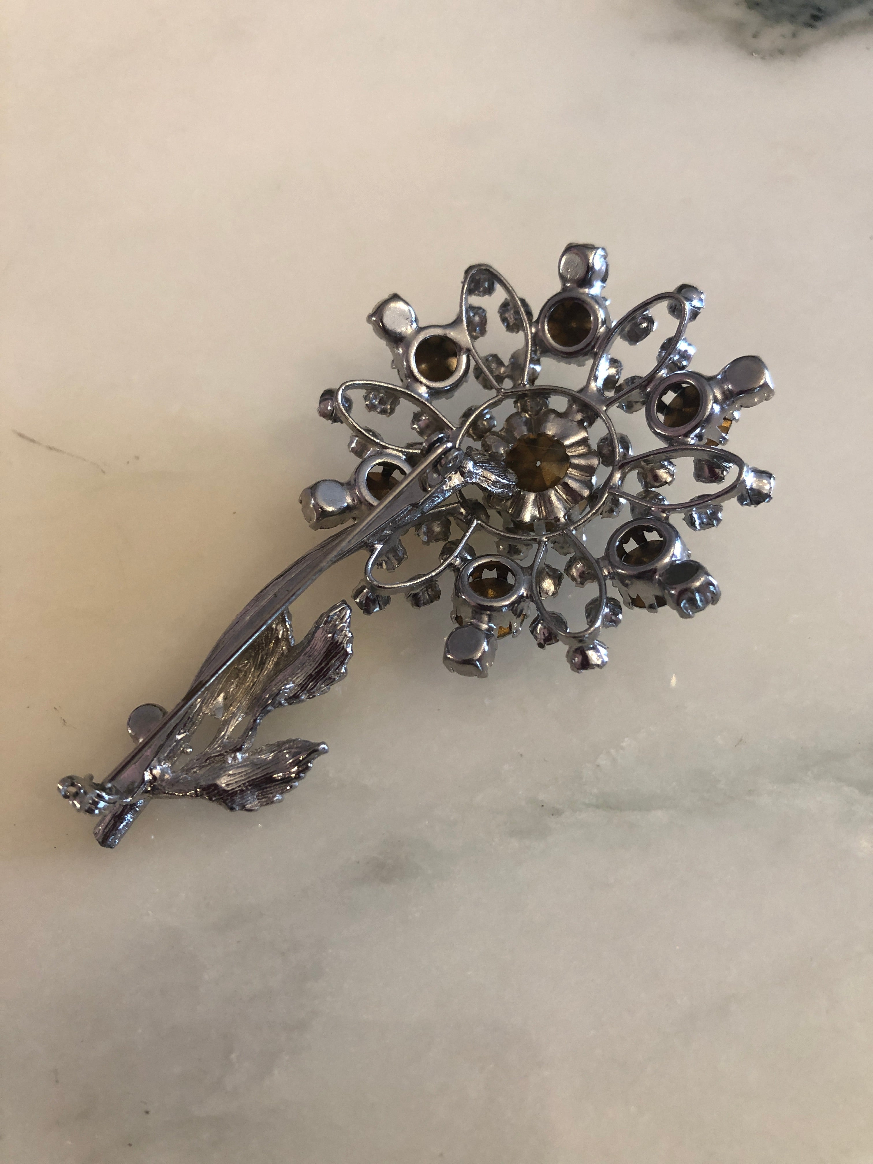A 7.5cm x 4.5cm vintage silver-tone crystal floral spray brooch with a graduating clear round crystal rhinestone flower head on fine wire stems and a textured silver-tone leaf and stem base, photographed on a white marble surface.