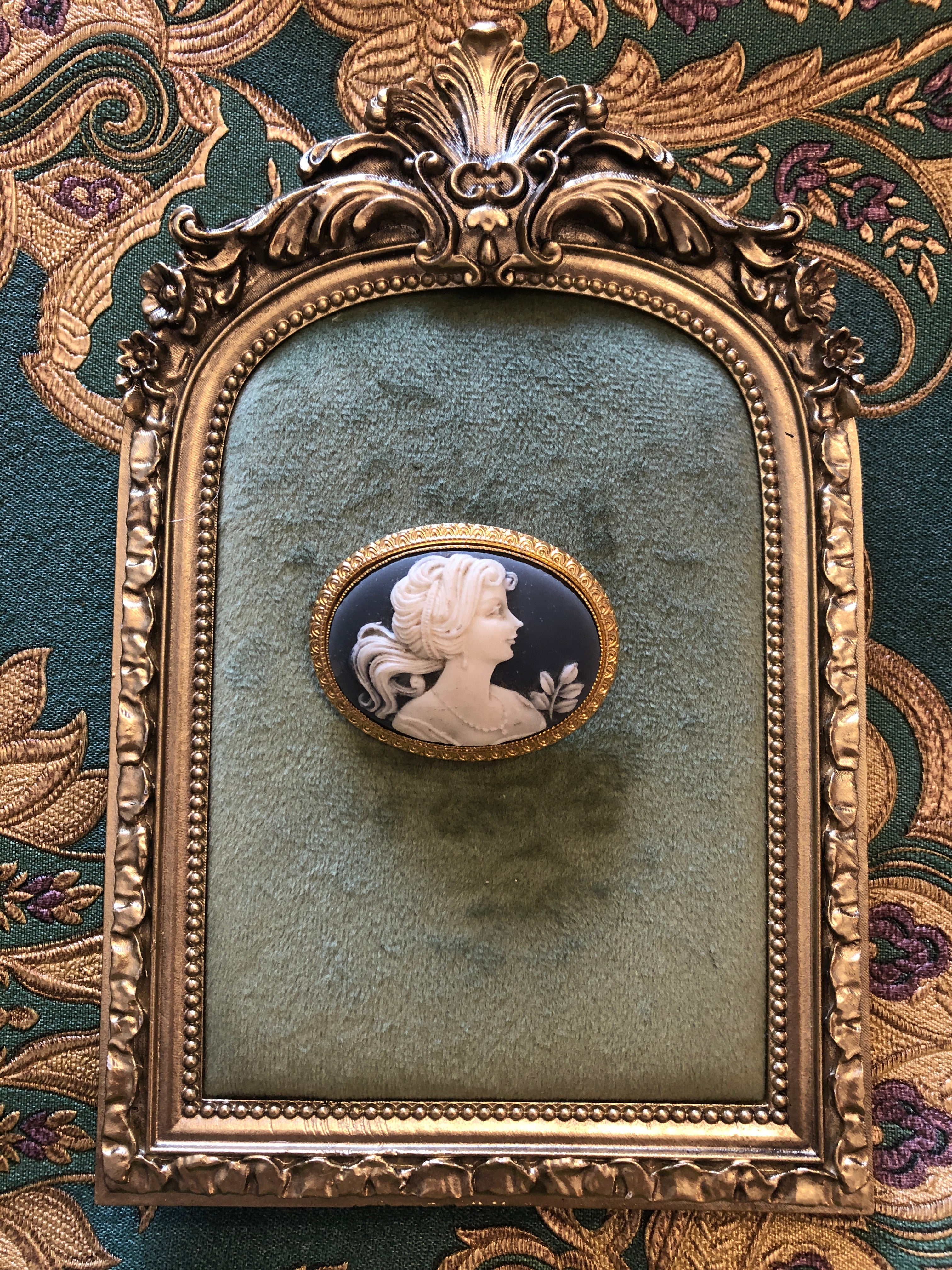 A 3.5cm x 4.5cm vintage cameo brooch with a carved white classical portrait on a deep blue ground, with flowing hair and flower bud detail, set in a gold-tone scrollwork frame. Photographed on a green velvet tray, with an ornate gold frame. The background is green and gold damask wallpaper