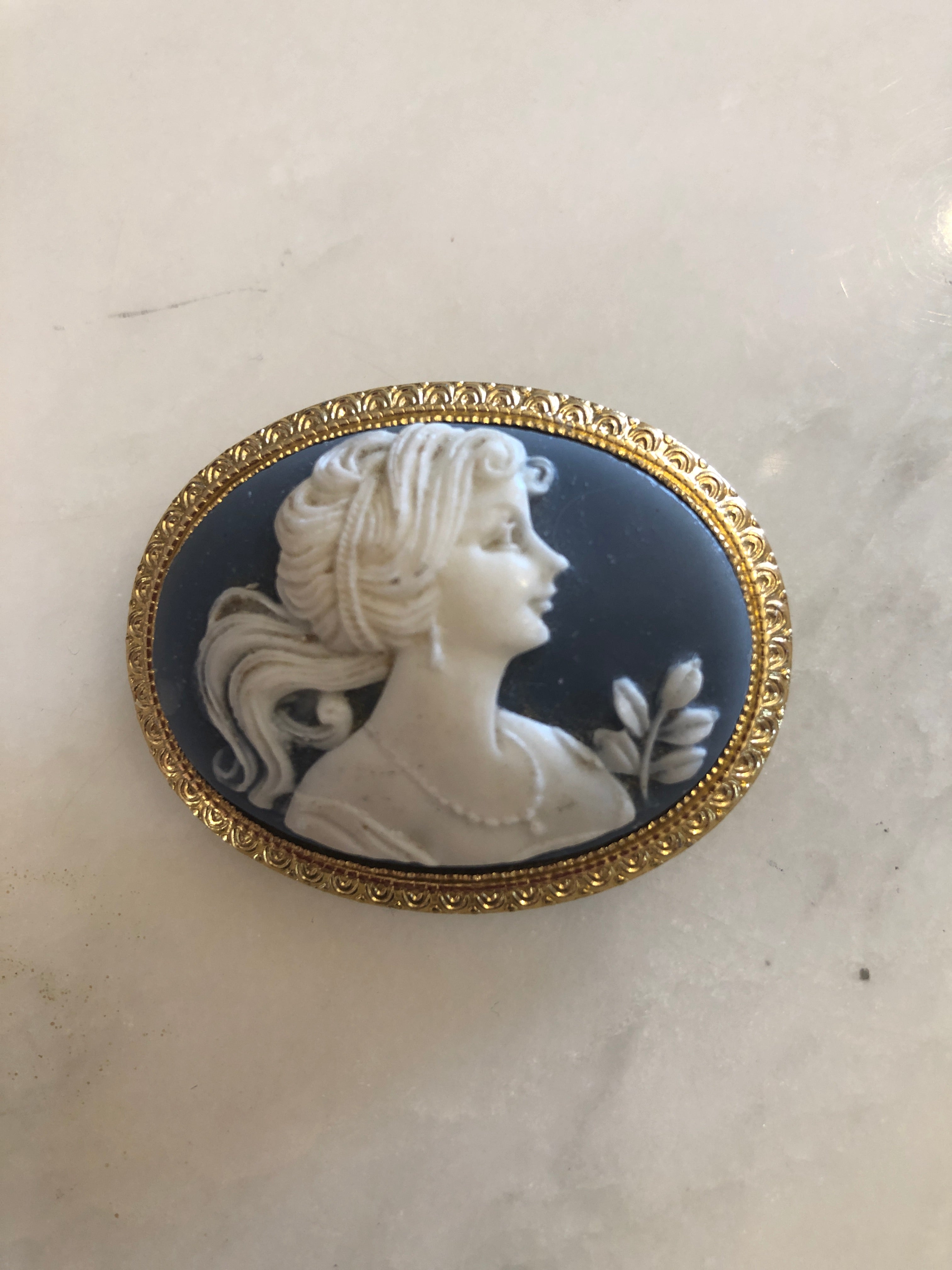 A 3.5cm x 4.5cm vintage cameo brooch with a carved white classical portrait on a deep blue ground, with flowing hair and flower bud detail, set in a gold-tone scrollwork frame, photographed on a white marble surface.