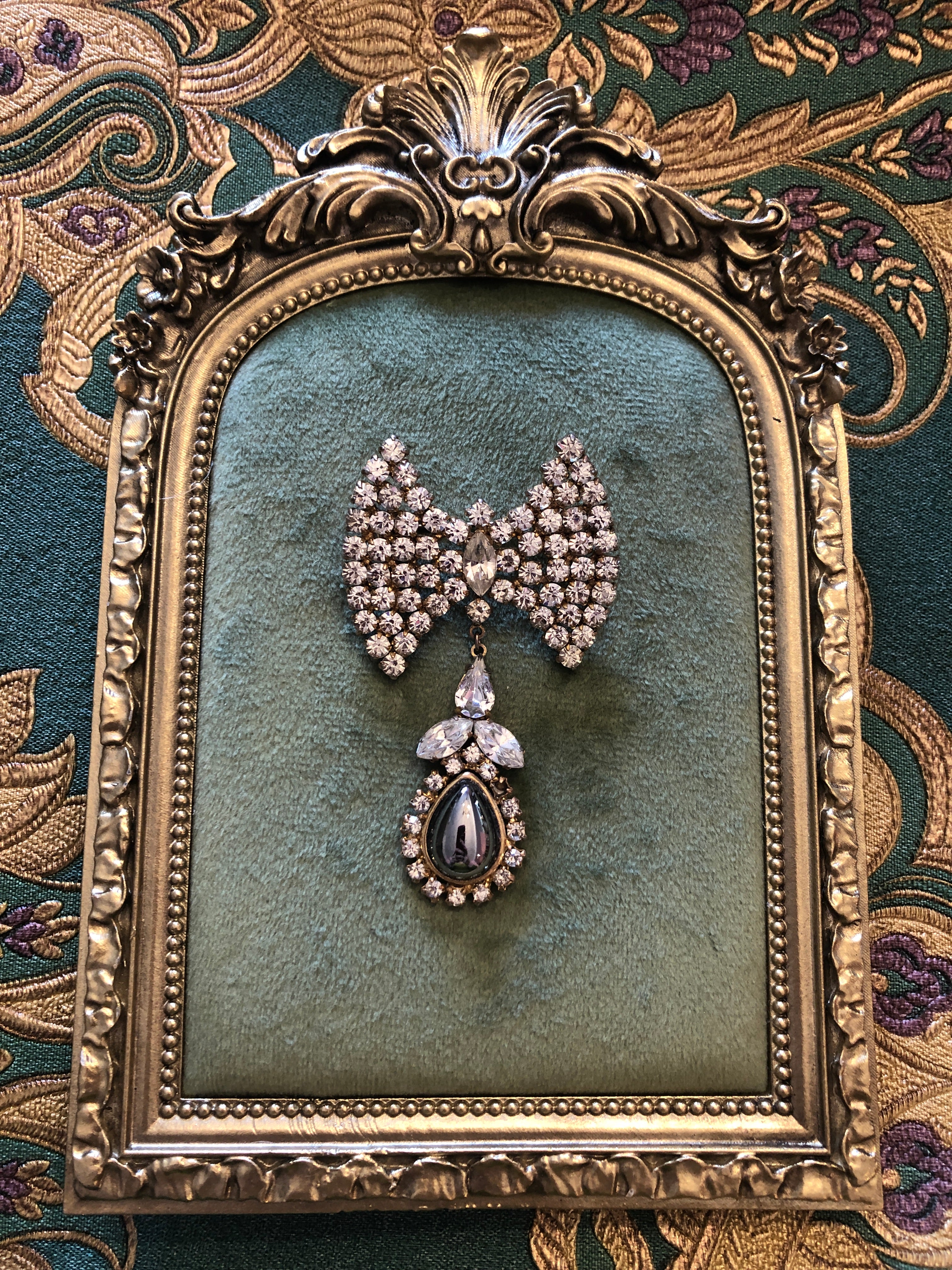 A 6.5cm x 4cm vintage gold-tone bow brooch densely pavé-set with clear crystal rhinestones, with a suspended drop of clear pear and marquise-cut crystals leading to a silver-tone teardrop cabochon. Photographed on a green velvet tray, with an ornate gold frame. The background is green and gold damask wallpaper