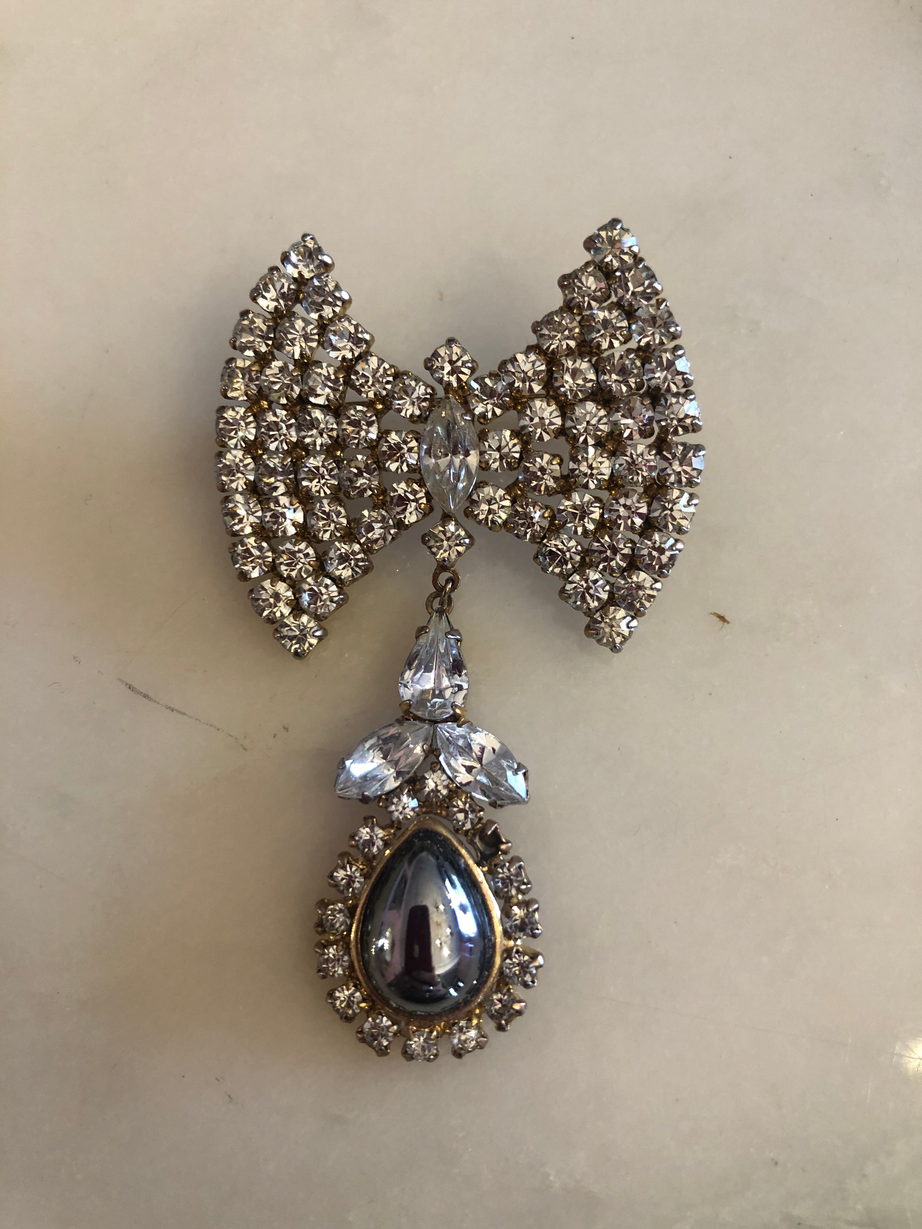 A 6.5cm x 4cm vintage gold-tone bow brooch densely pavé-set with clear crystal rhinestones, with a suspended drop of clear pear and marquise-cut crystals leading to a silver-tone teardrop cabochon photographed on a white marble surface.