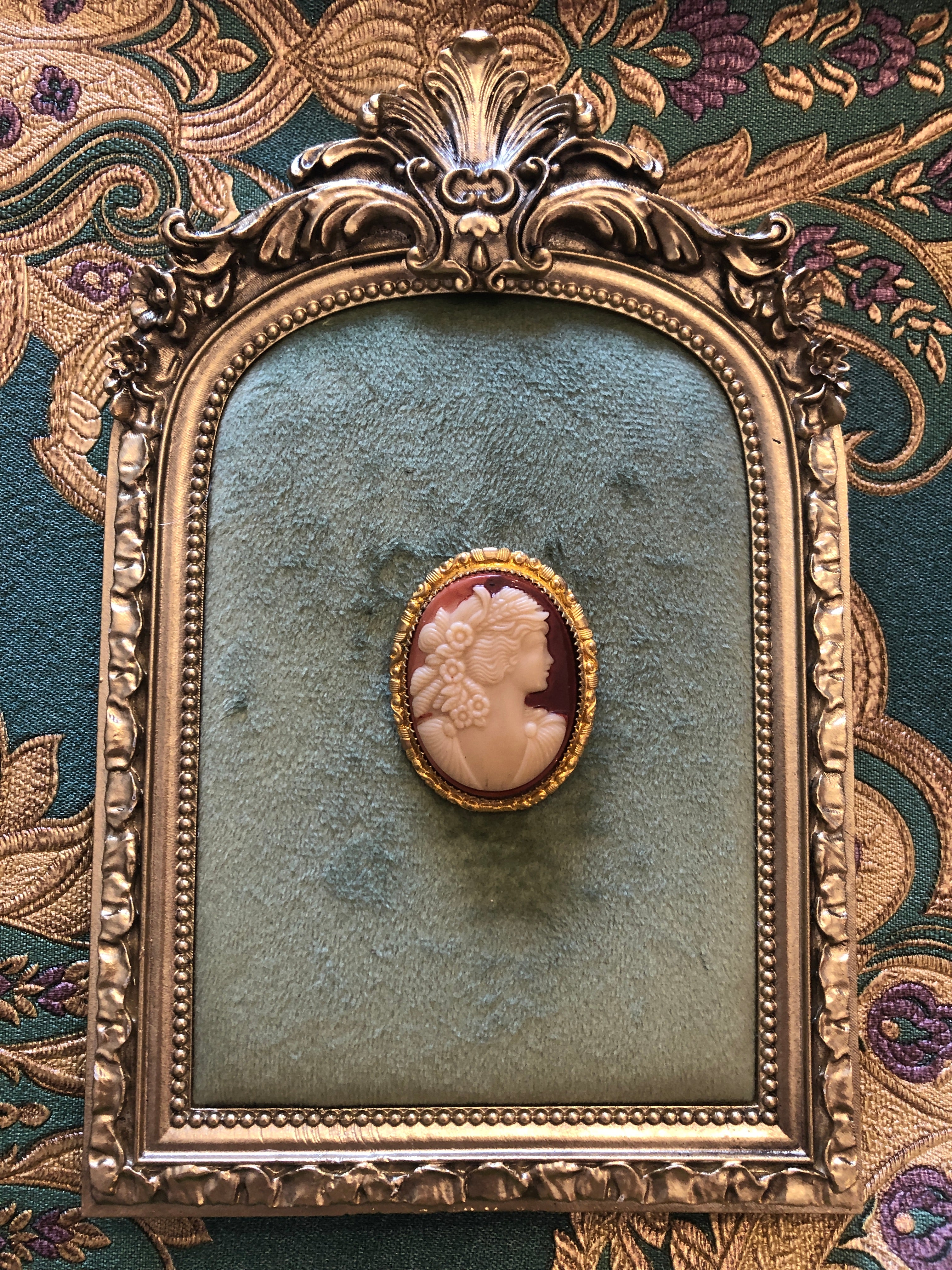 A 4cm x 3cm vintage shell cameo brooch with a carved cream relief portrait of a flower-crowned goddess in profile on a pink-mauve ground, set in a gold-tone scrollwork frame. Photographed on a green velvet tray with an ornate gold frame. The background is green and gold damask wallpaper