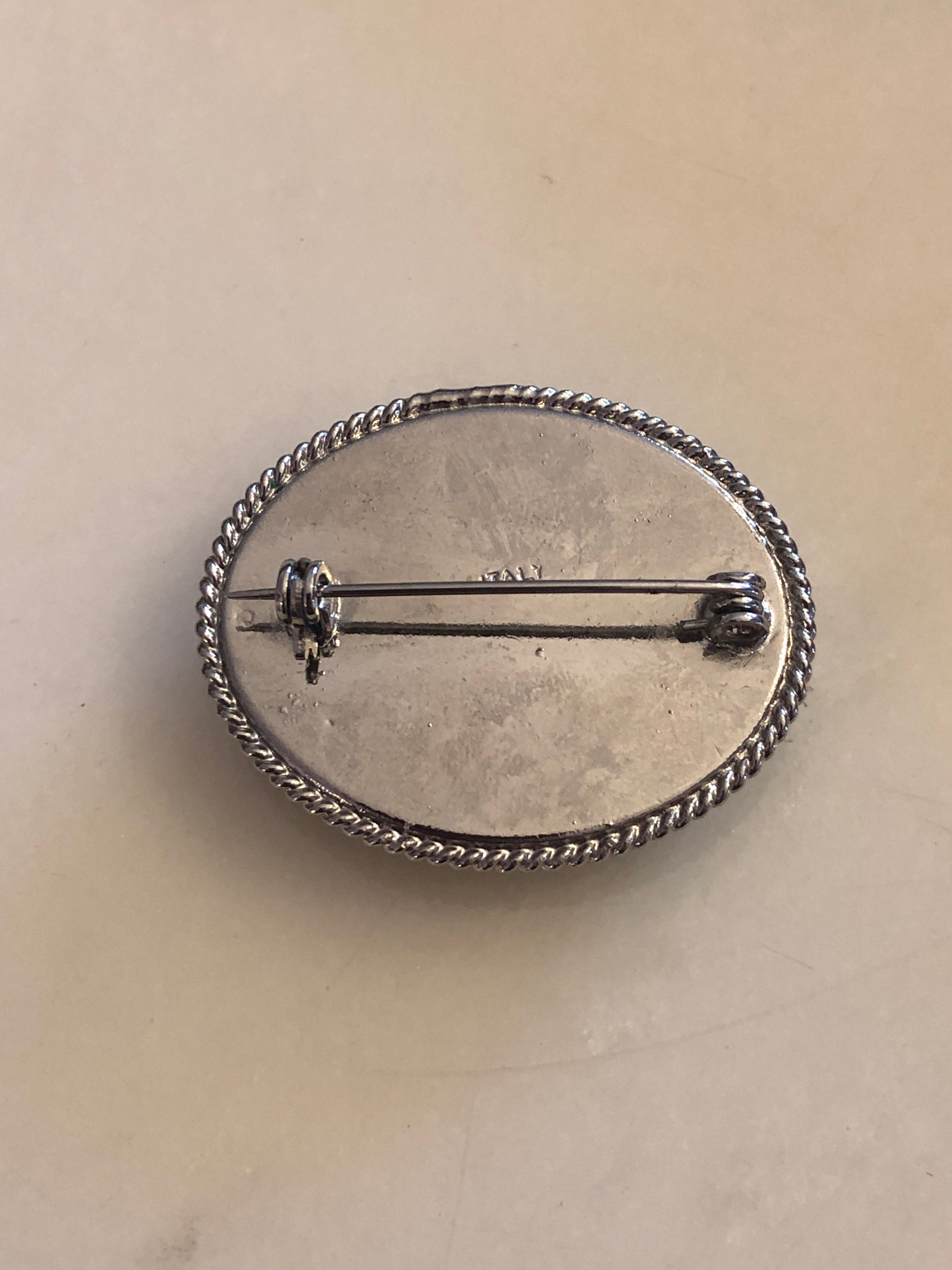 A 3cm x 4cm vintage Italian micro mosaic oval brooch featuring a hand assembled glass tessera design of forget-me-not flowers in cornflower blue, white and yellow on a blue ground, set within a silver-tone frame with twisted rope edge detail, photographed on a white marble surface.