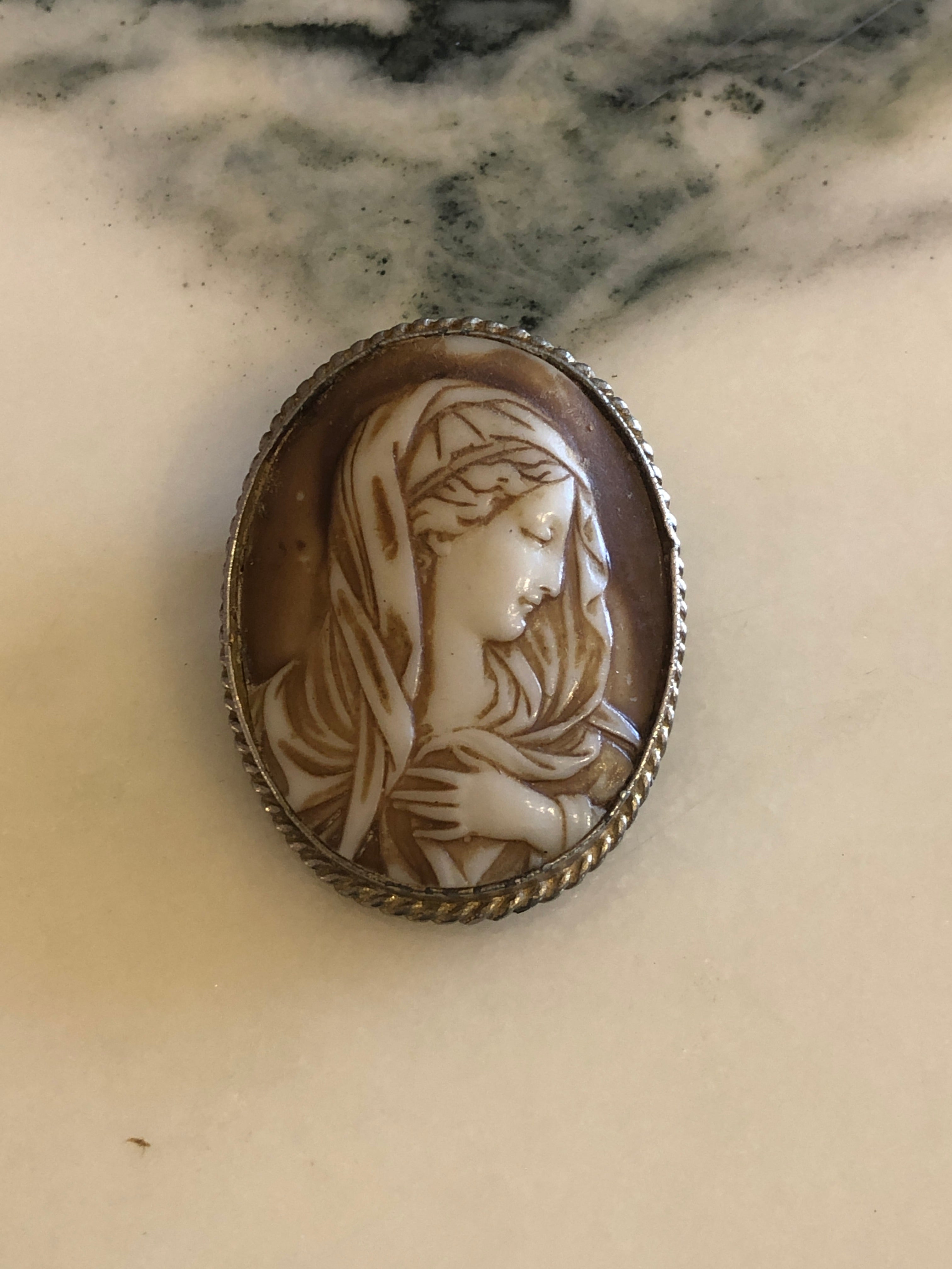  5cm x 4cm vintage signed Cordia cameo brooch with a carved white relief Madonna portrait with bowed head and clasped hands on a warm brown ground, in a silver-tone twisted rope frame, photographed on a white marble surface.