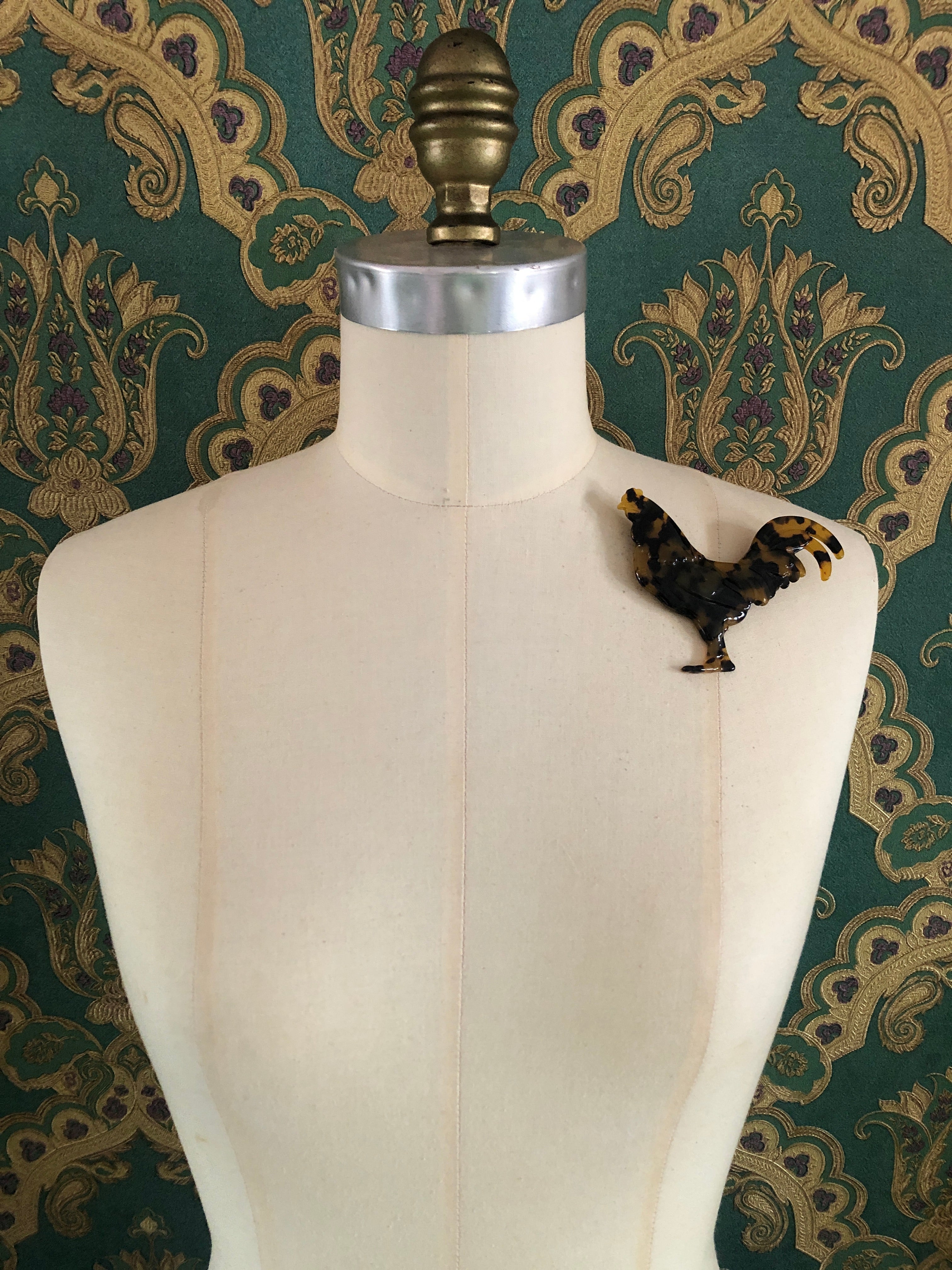 A Broochella Animalia Collection rooster brooch in brown and amber tortoiseshell Italian cellulose acetate with sculpted feather and tail detail measuring 8cm x 10cm, photographed on a white marble surface.
