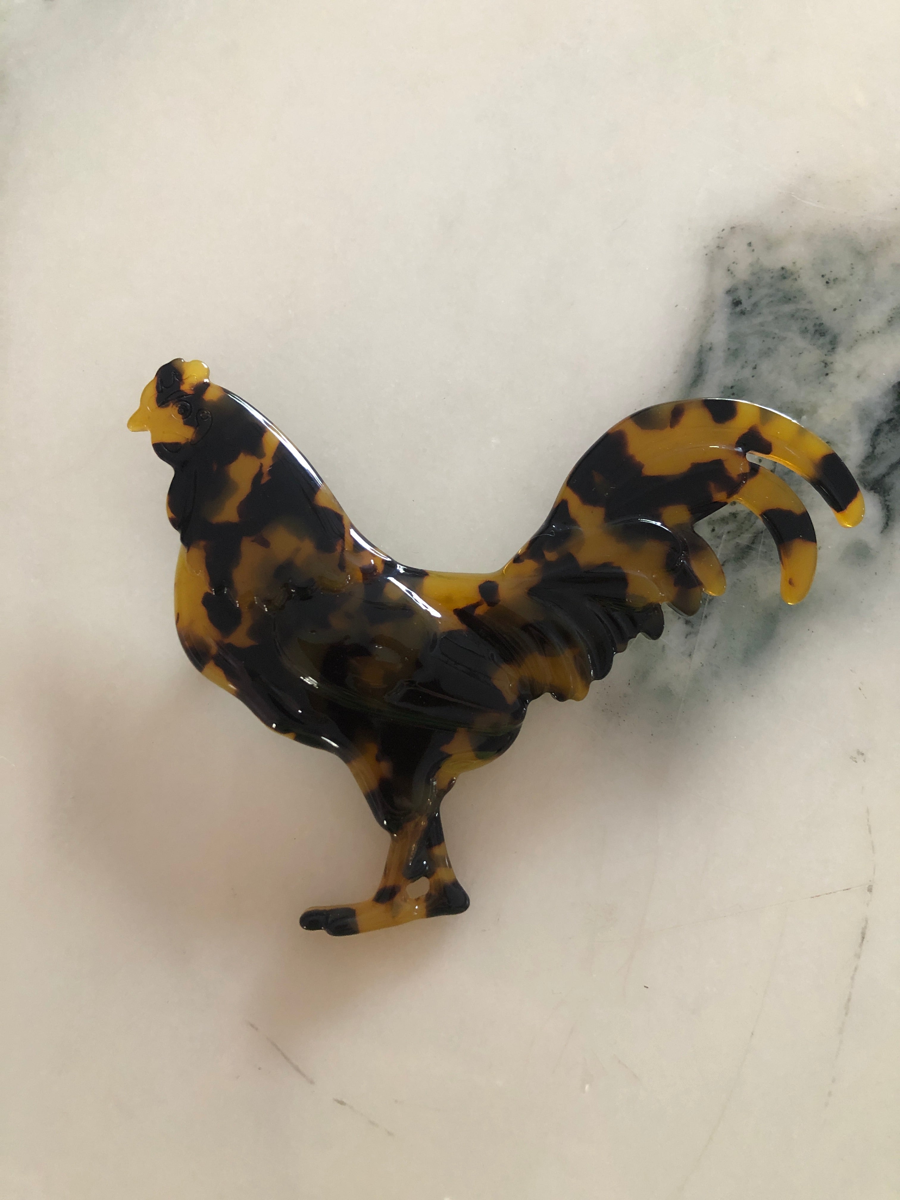 A Broochella Animalia Collection rooster brooch in brown and amber tortoiseshell Italian cellulose acetate with sculpted feather and tail detail measuring 8cm x 10cm, photographed on a white marble surface.
