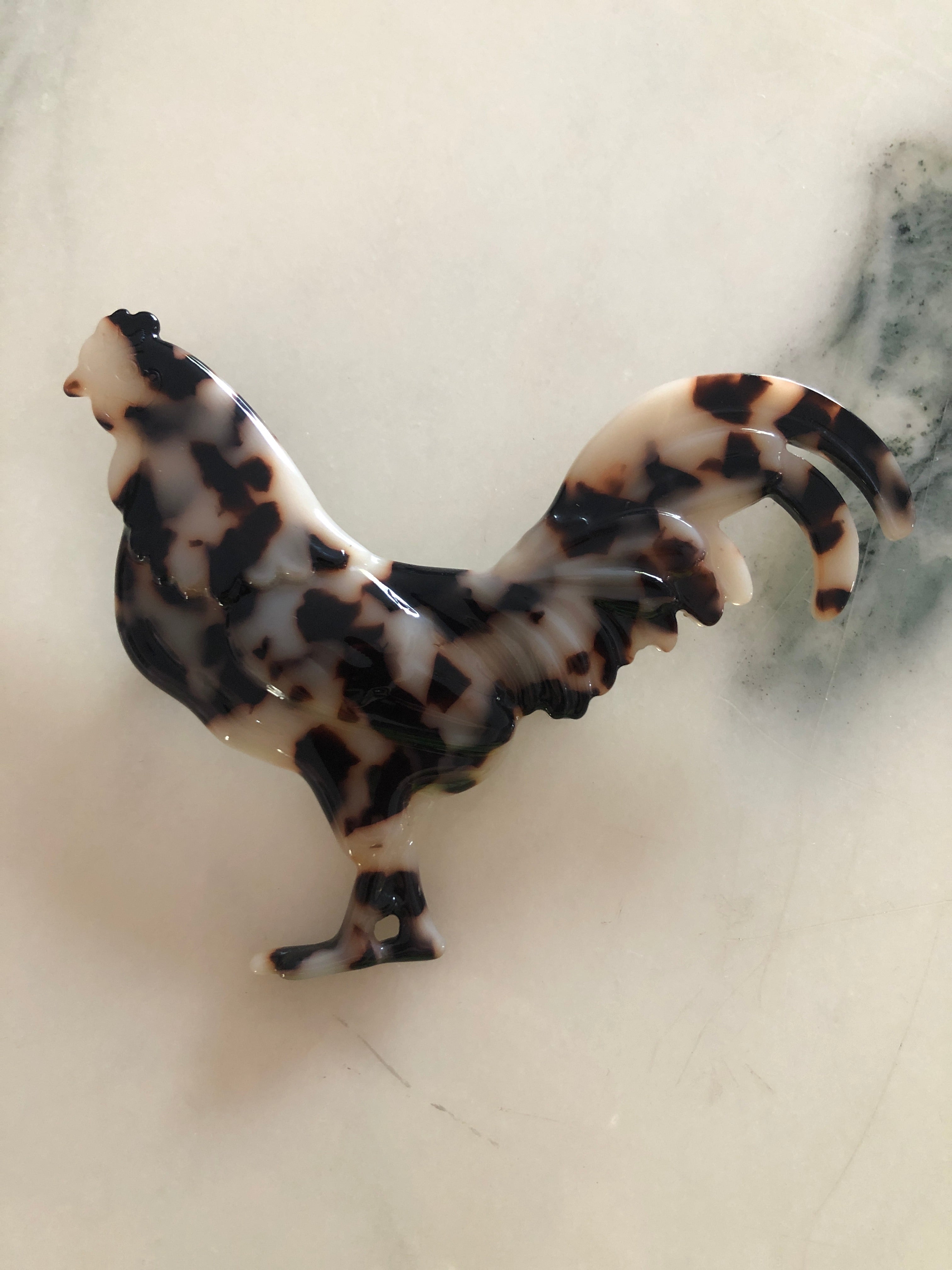 A Broochella Animalia Collection rooster brooch in brown and amber tortoiseshell Italian cellulose acetate with sculpted feather and tail detail measuring 8cm x 10cm, photographed on a white marble surface.
