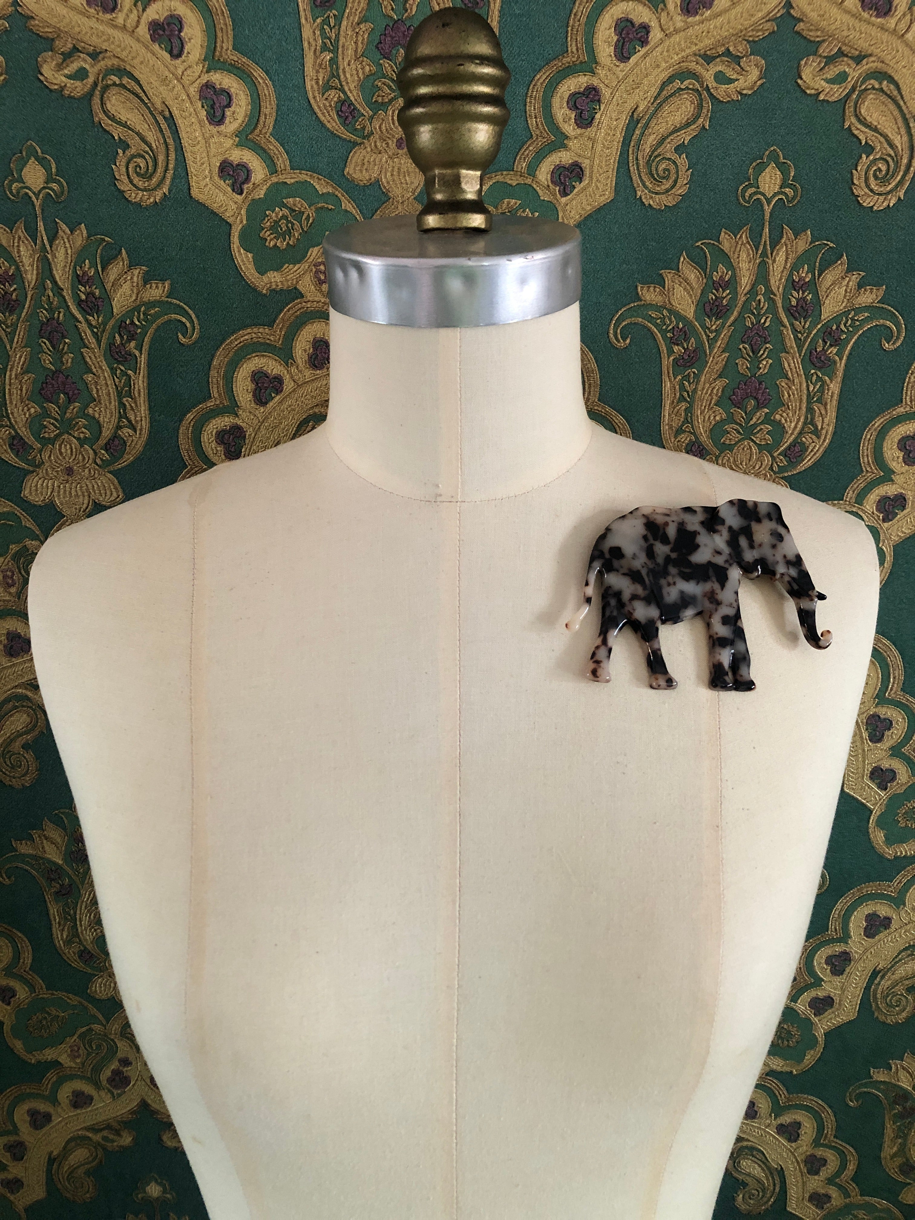 A Broochella Animalia Collection elephant brooch in black and white tortoiseshell Italian cellulose acetate with sculpted curled trunk detail measuring 8cm x 11cm, photographed on a white marble surface.