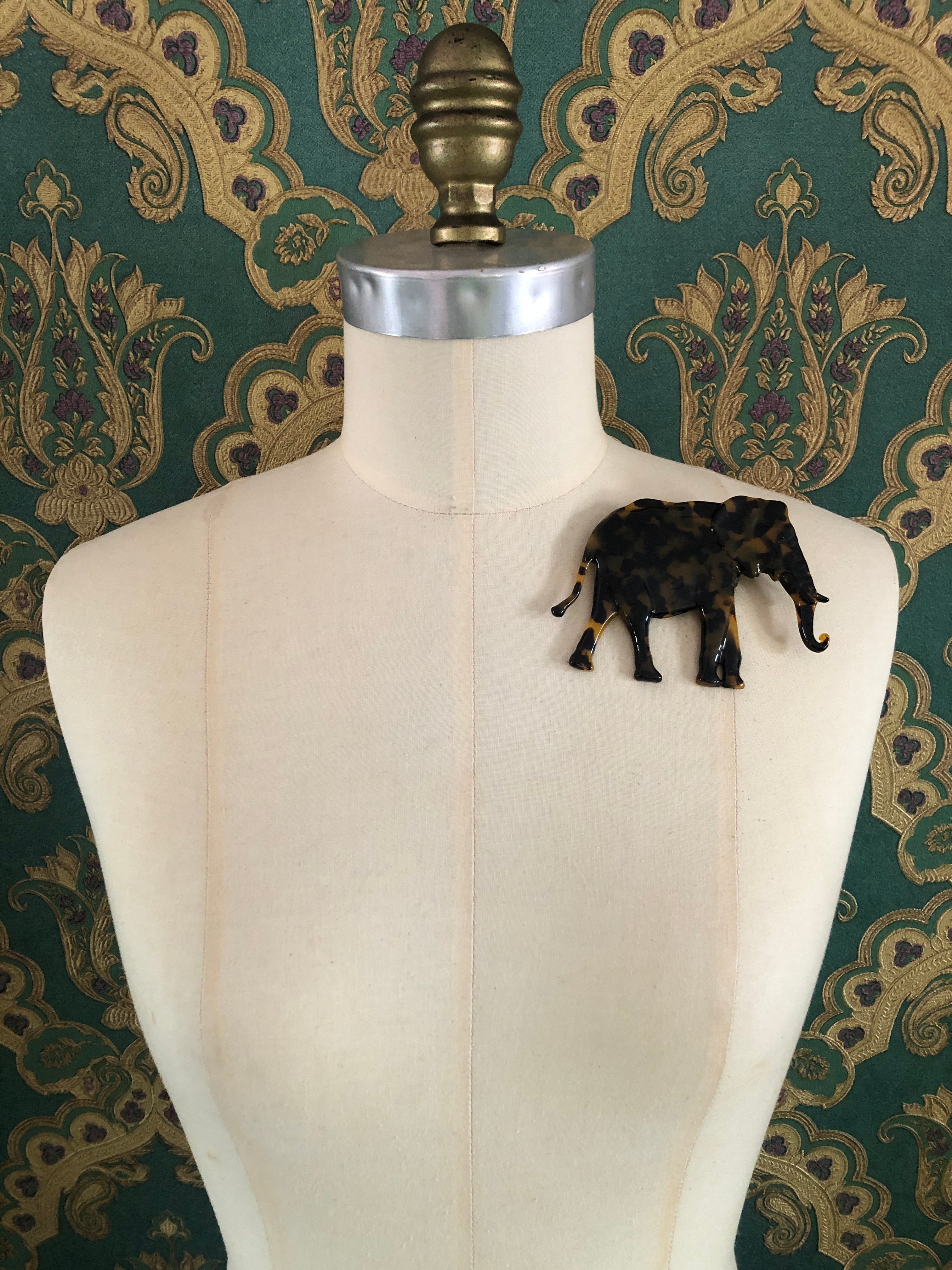 A Broochella Animalia Collection elephant brooch in black and white tortoiseshell Italian cellulose acetate with sculpted curled trunk detail measuring 8cm x 11cm, photographed on a white marble surface.