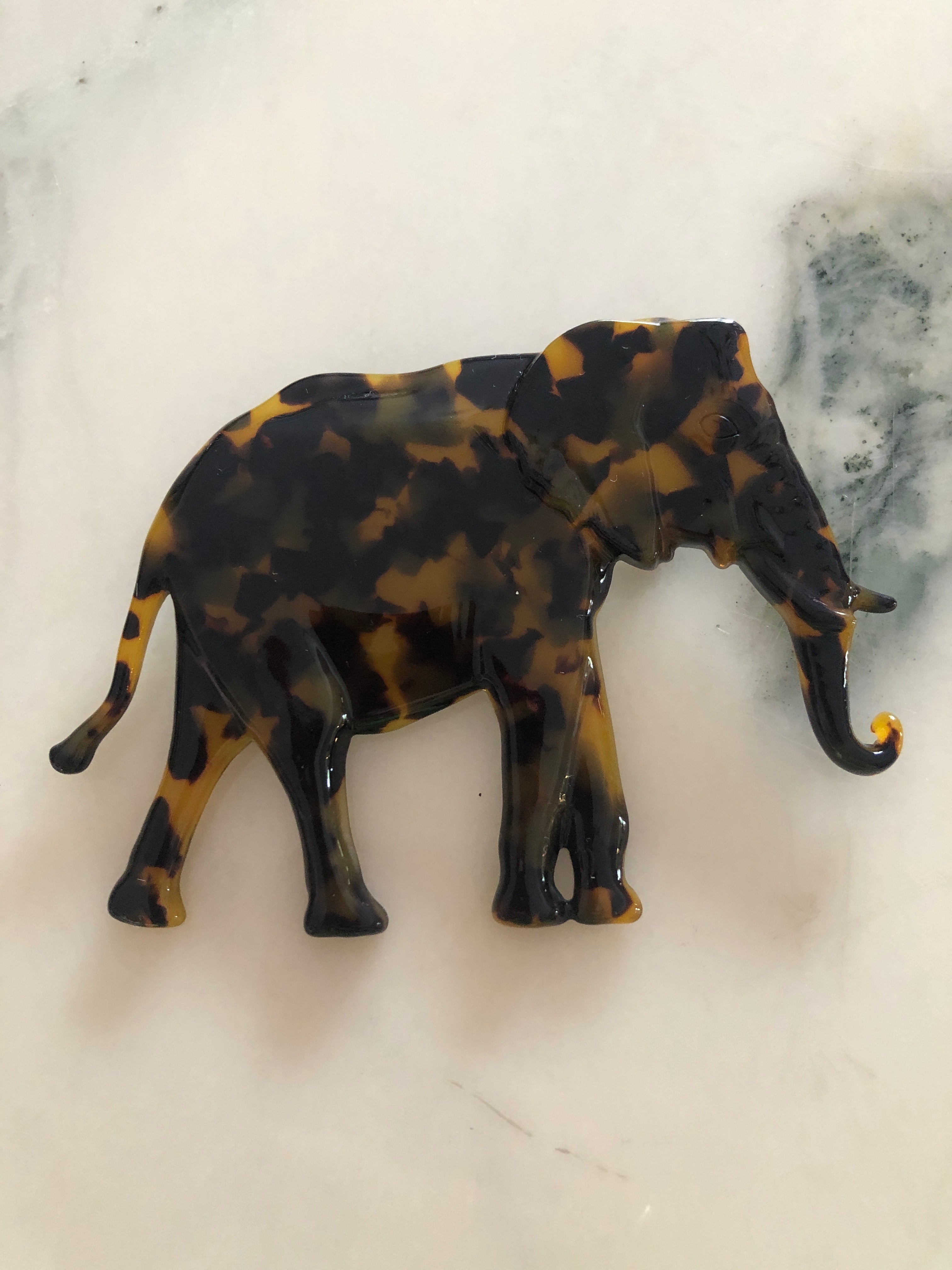 A Broochella Animalia Collection elephant brooch in black and white tortoiseshell Italian cellulose acetate with sculpted curled trunk detail measuring 8cm x 11cm, photographed on a white marble surface.