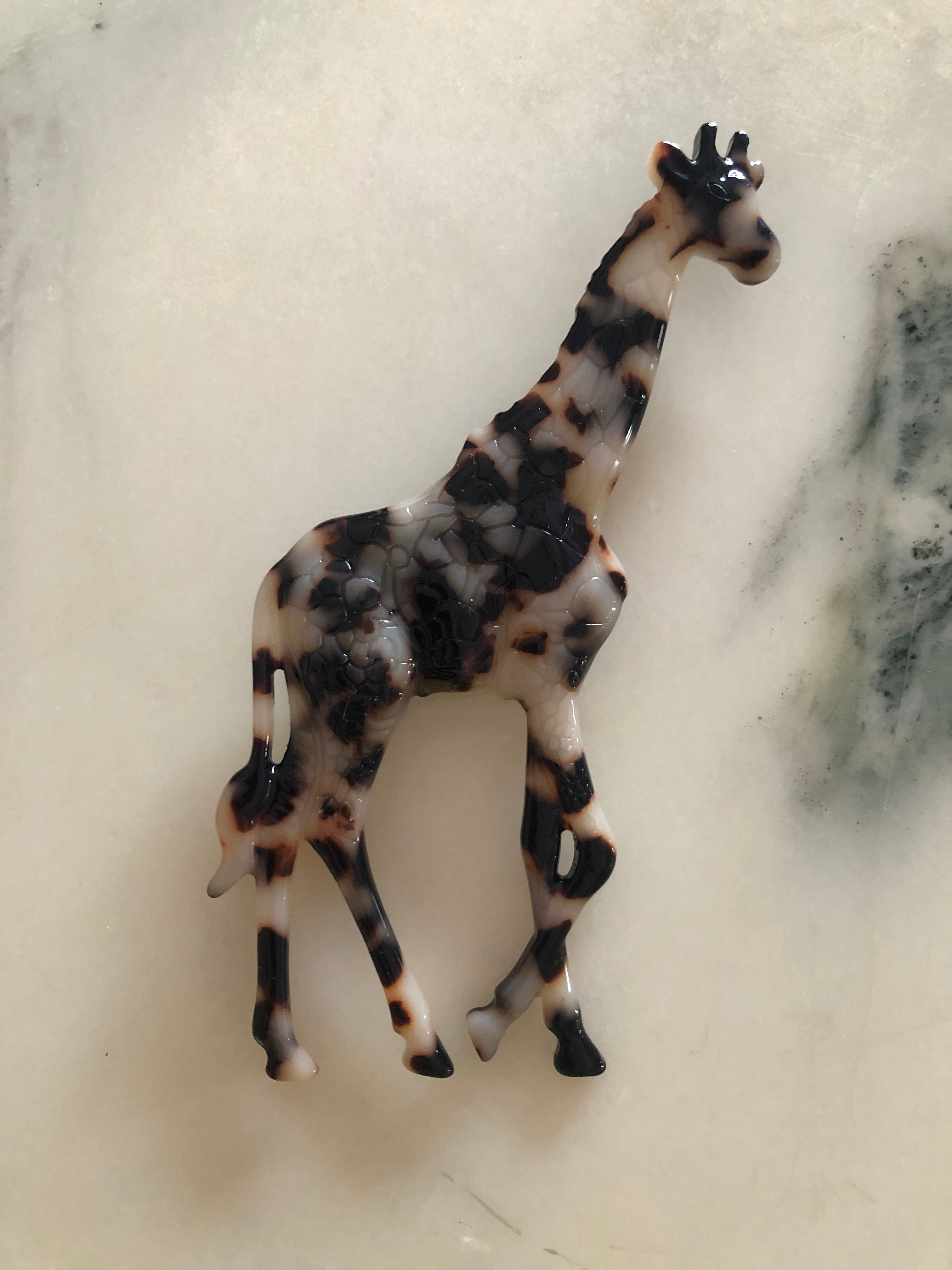 A Broochella Animalia Collection standing giraffe brooch in black and white tortoiseshell Italian cellulose acetate with sculpted spot patterning measuring 12cm x 7cm, photographed on a white marble surface.