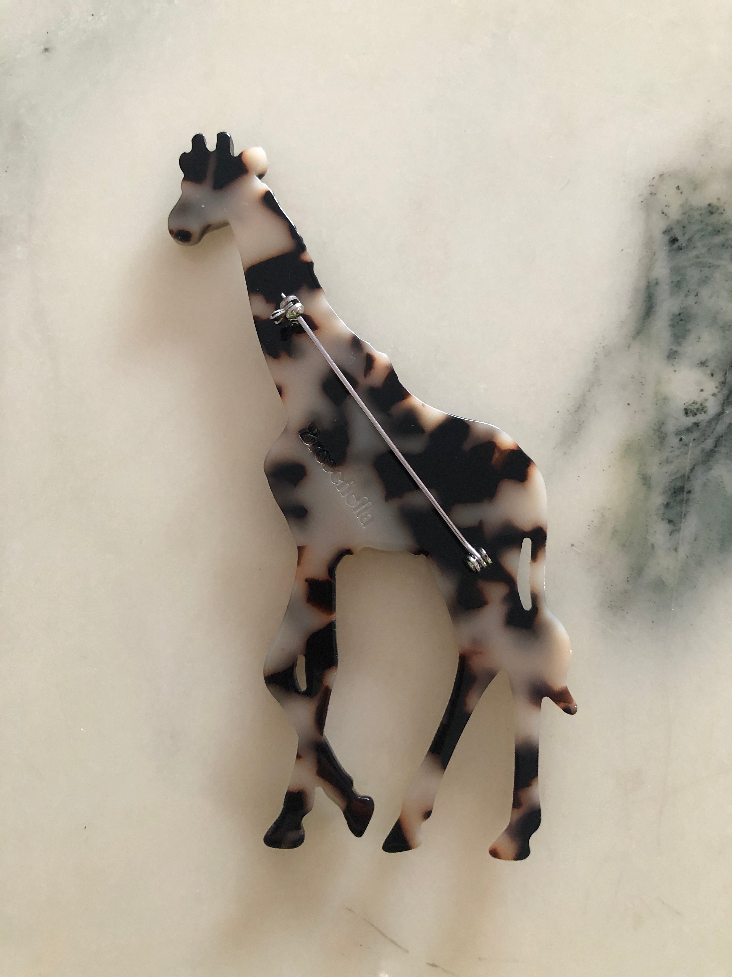 A Broochella Animalia Collection giraffe brooch and men's lapel pin in brown and beige tortoiseshell Italian cellulose acetate with sculpted spot patterning measuring 12cm x 7cm, photographed on a white marble surface.