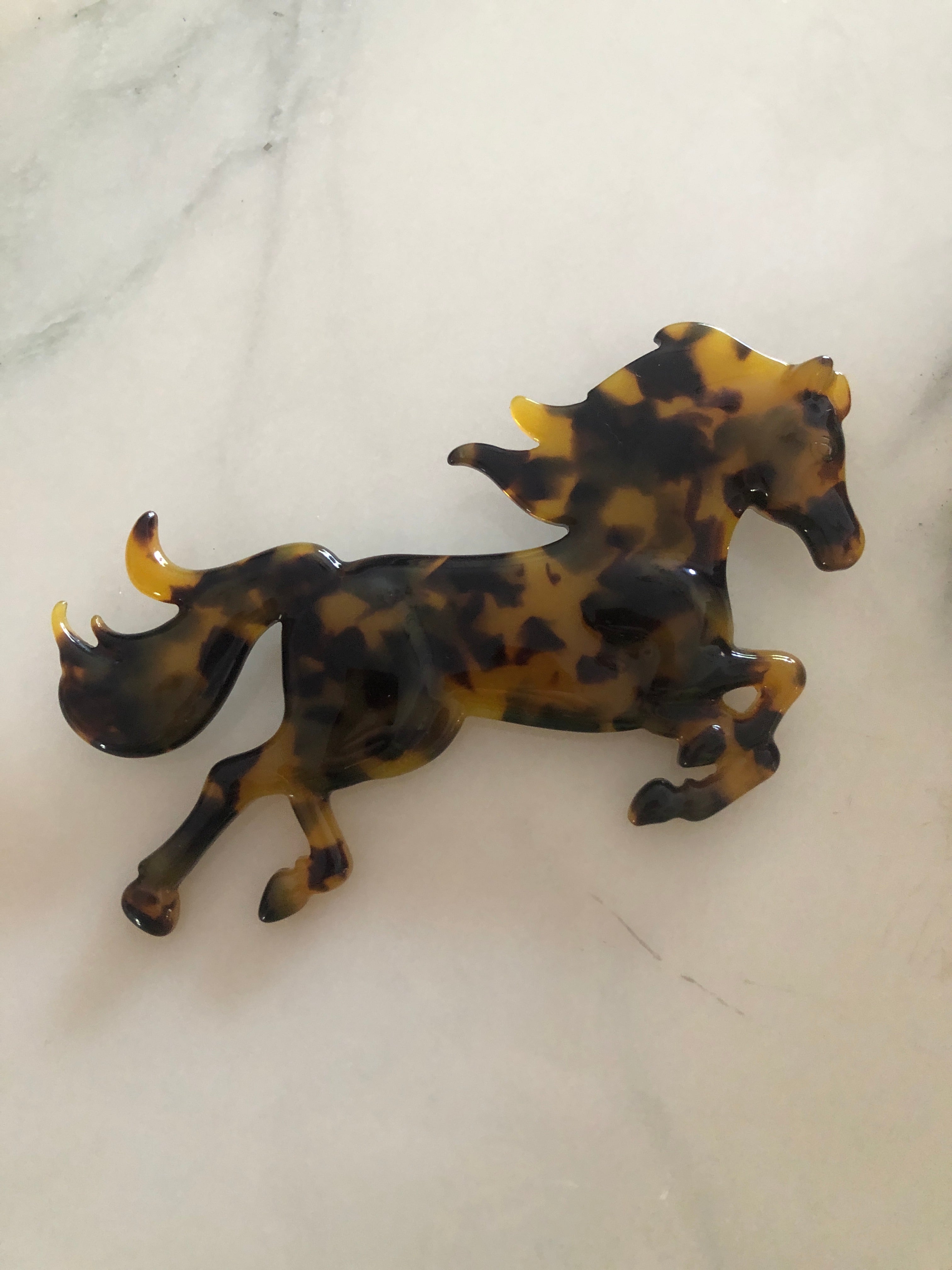 A Broochella Animalia Collection running horse brooch in Italian tortoiseshell cellulose acetate measuring 9cm x 11cm, photographed on a white marble surface.