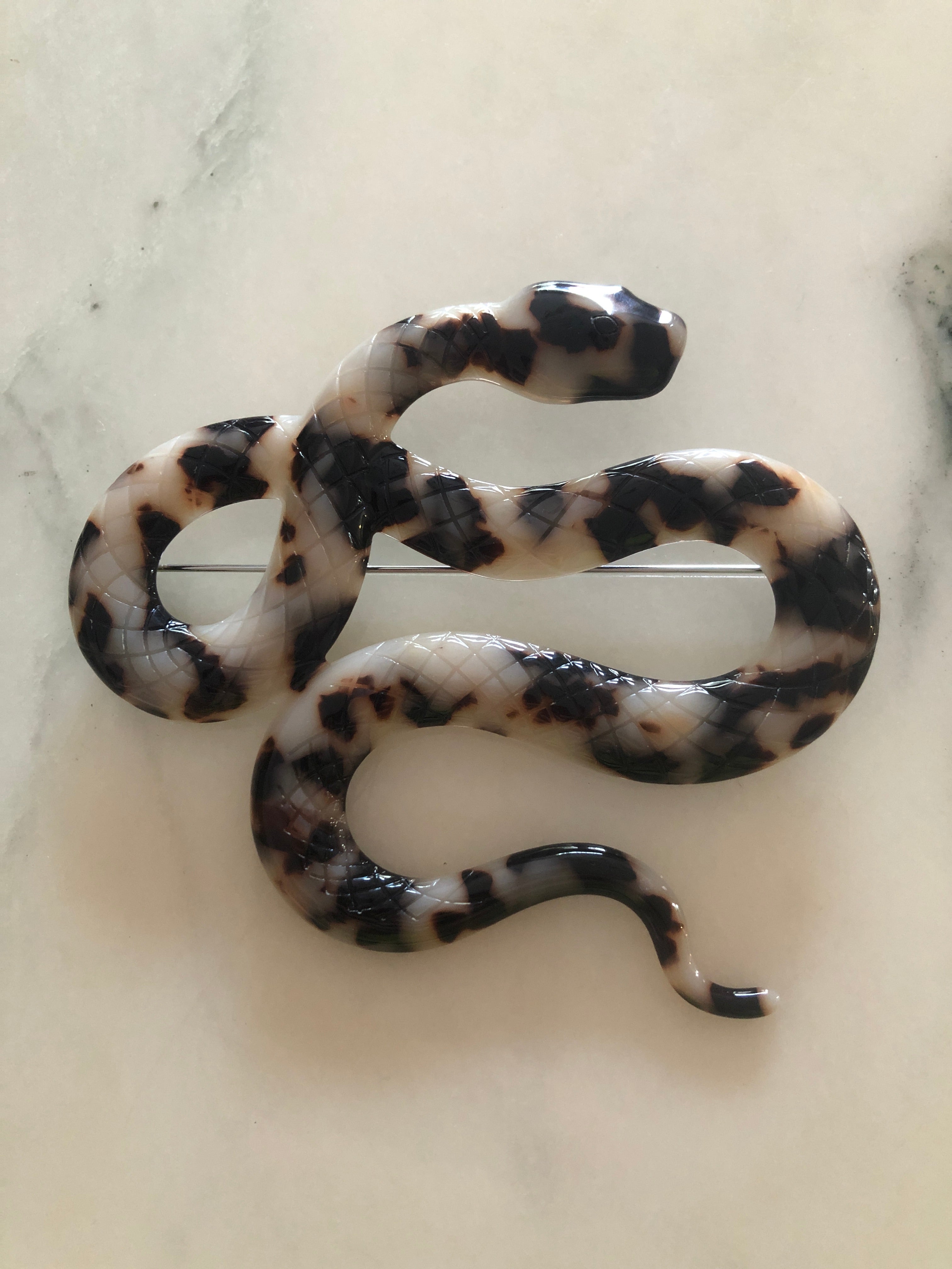 A Broochella Animalia Collection coiled snake brooch in black and white tortoiseshell Italian cellulose acetate with detailed scale texture measuring 9cm x 10cm, photographed on a white marble surface.