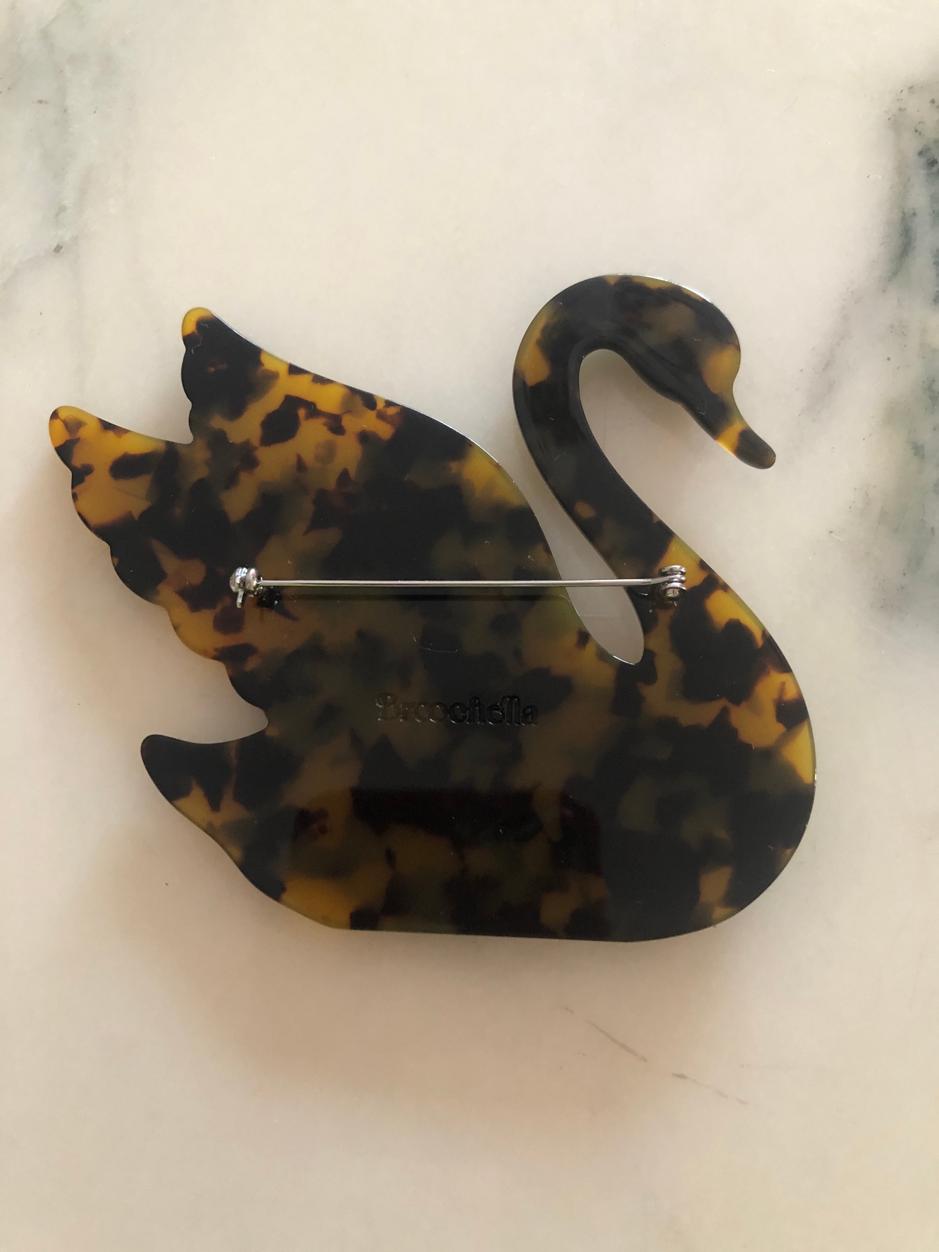 A Broochella Animalia Collection beige tortoiseshell swan brooch and men's lapel pin in Italian cellulose acetate. The background is white marble