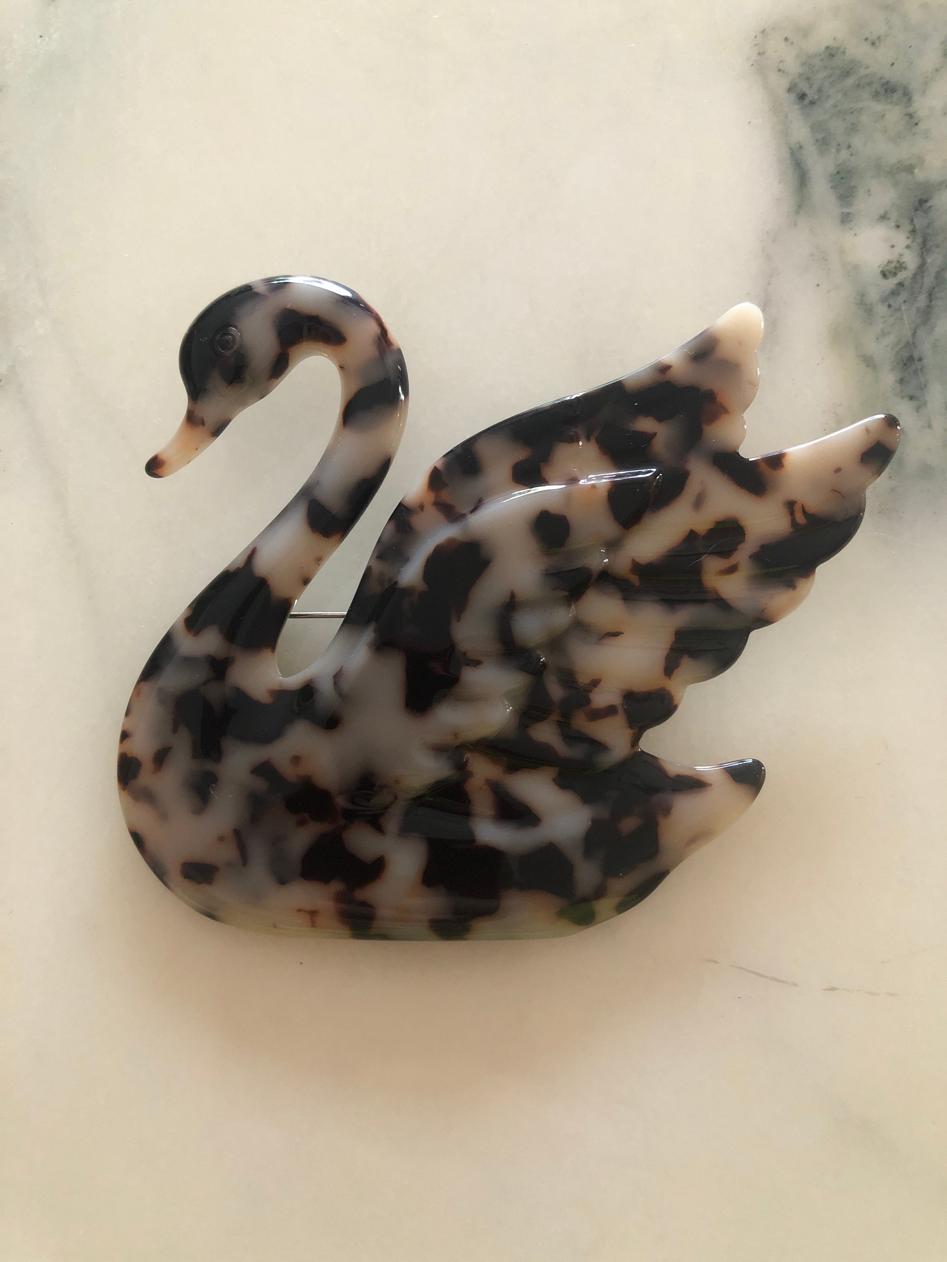 A Broochella Animalia Collection beige tortoiseshell swan brooch and men's lapel pin in Italian cellulose acetate. The background is white marble