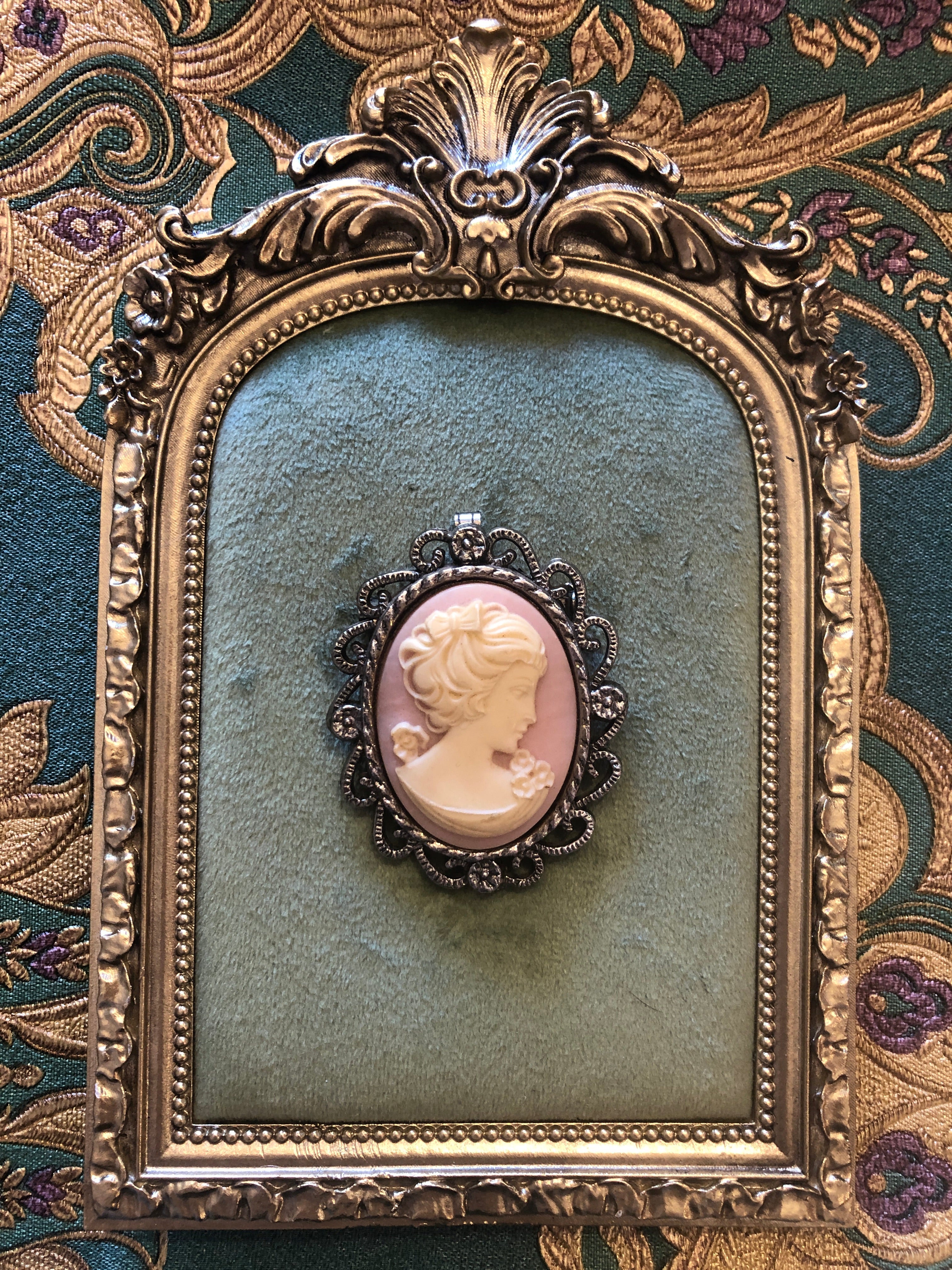 A 5.5cm x 4.5cm vintage cameo brooch featuring a carved cream classical portrait on a blush pink ground set within a silver-tone openwork filigree scrollwork frame, photographed on a white marble surface.