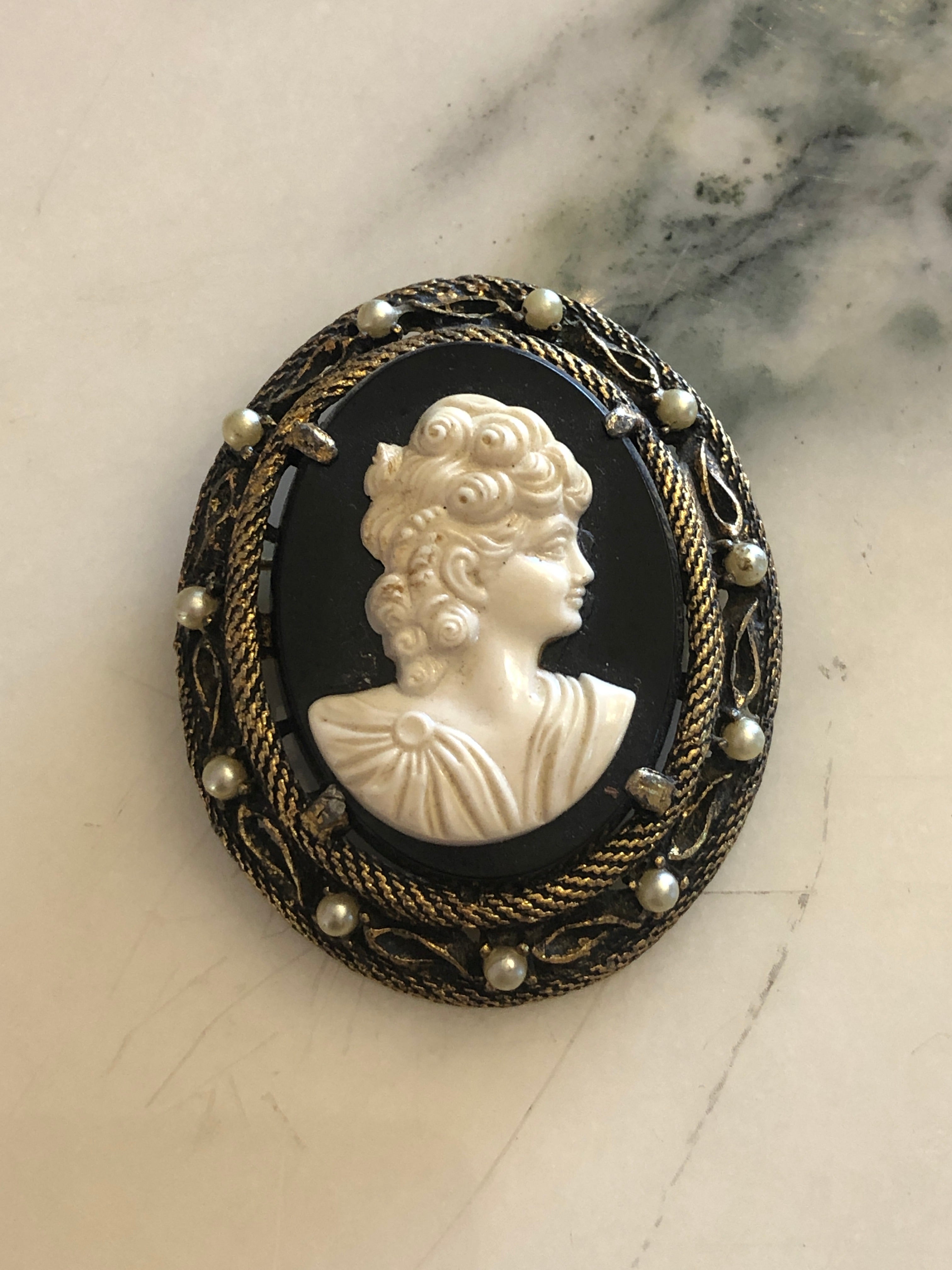 A 5.5cm x 4.5cm vintage cameo brooch featuring a carved white classical portrait on a deep black ground set within a gold-tone rope twist filigree frame with seed pearl accents, photographed on a white marble surface.
