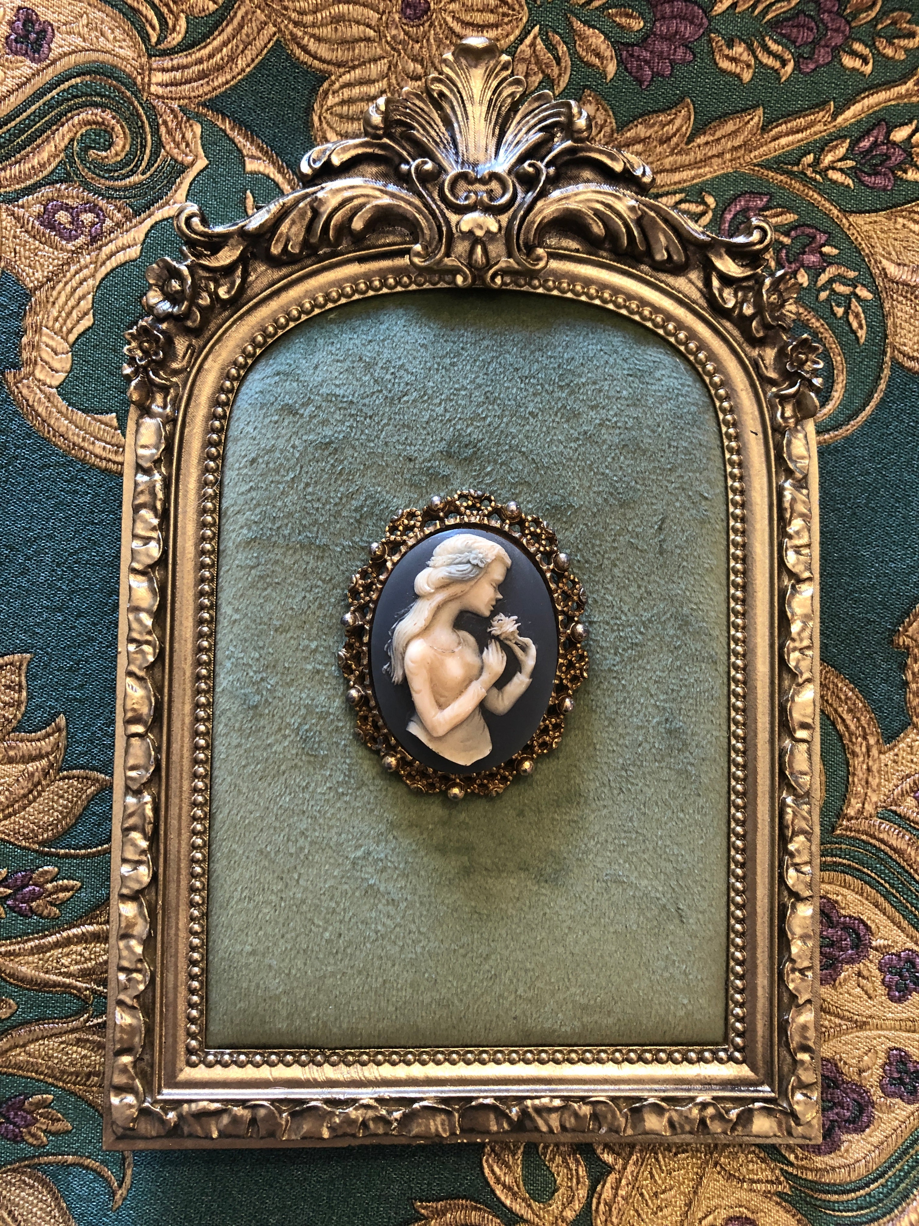 A 5cm x 4cm vintage cameo brooch featuring a carved white full figure classical portrait holding a floral bouquet on a deep slate blue ground, set within a gold-tone openwork filigree frame with silver bead accents, photographed on a white marble surface.
