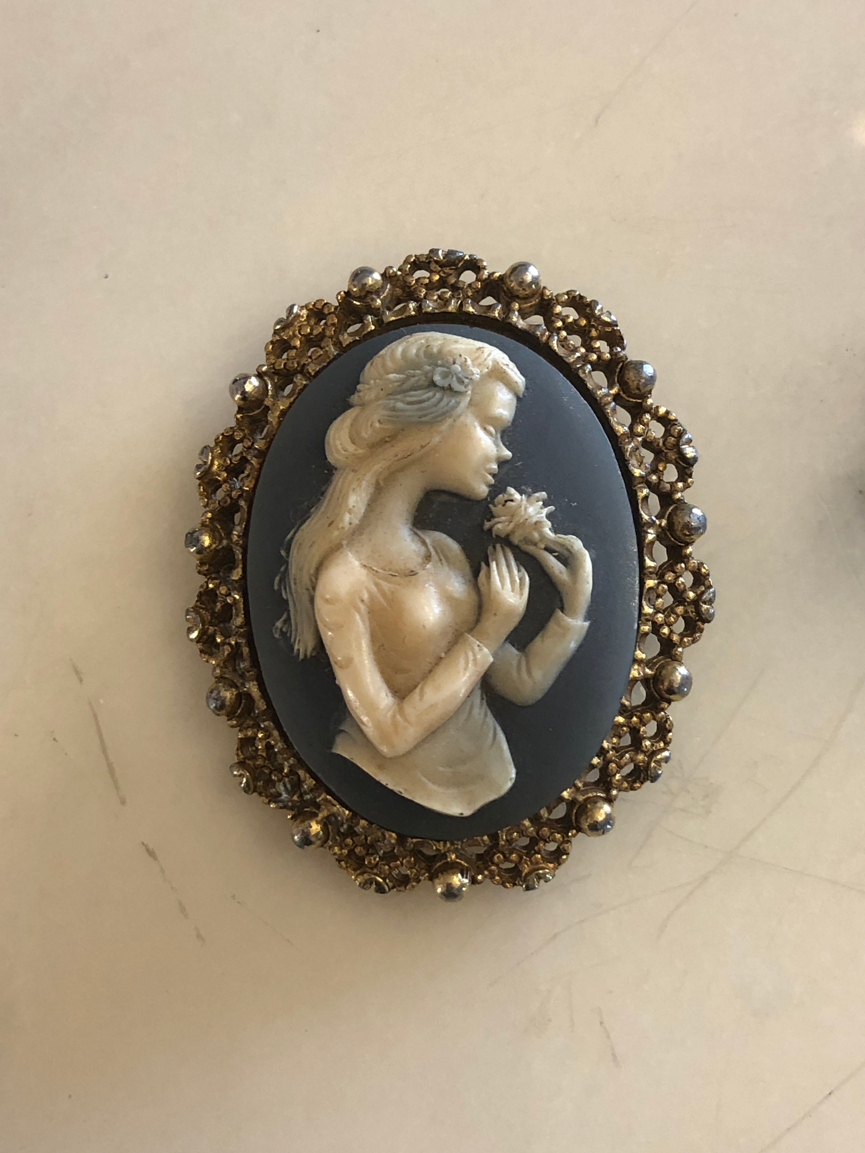 A 5cm x 4cm vintage cameo brooch featuring a carved white full figure classical portrait holding a floral bouquet on a deep slate blue ground, set within a gold-tone openwork filigree frame with silver bead accents, photographed on a white marble surface.